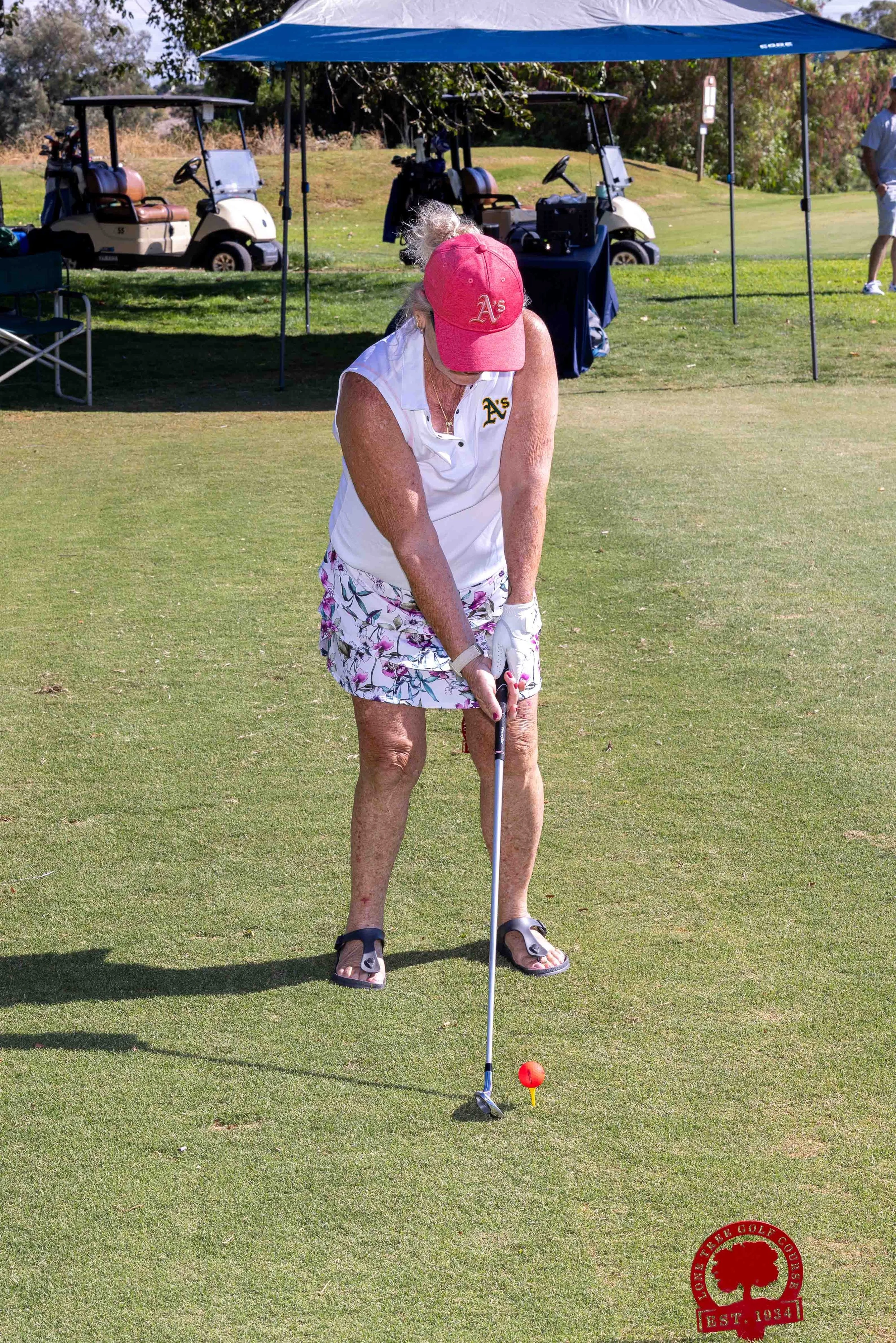 An elderly woman playing golf on a sunny day, wearing a red cap and floral shorts, preparing to putt on the green. There are golf carts in the background and a blue canopy providing shade.