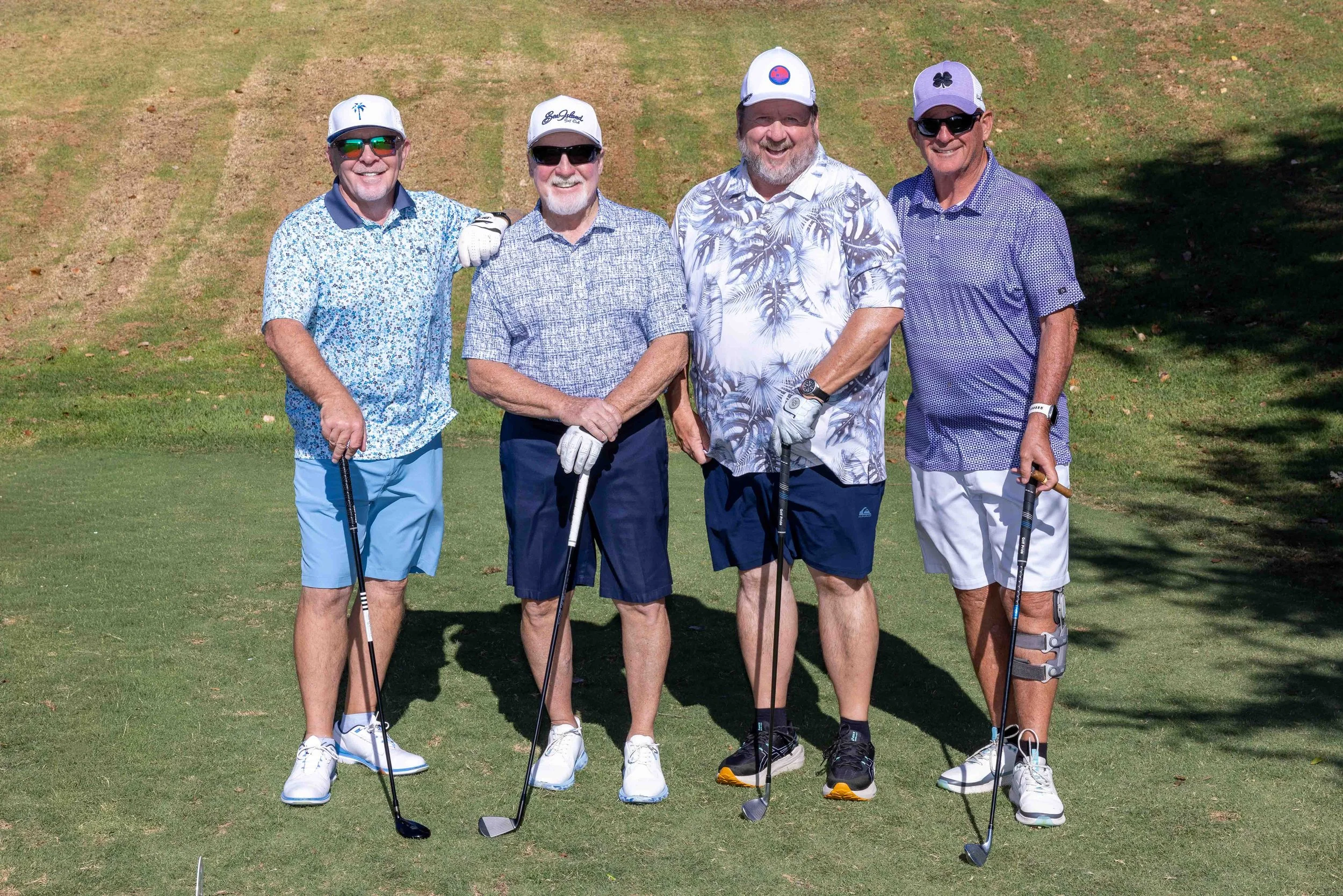 Four men standing together on a golf course, holding golf clubs, smiling, dressed in casual golf attire including hats, sunglasses, and shorts.