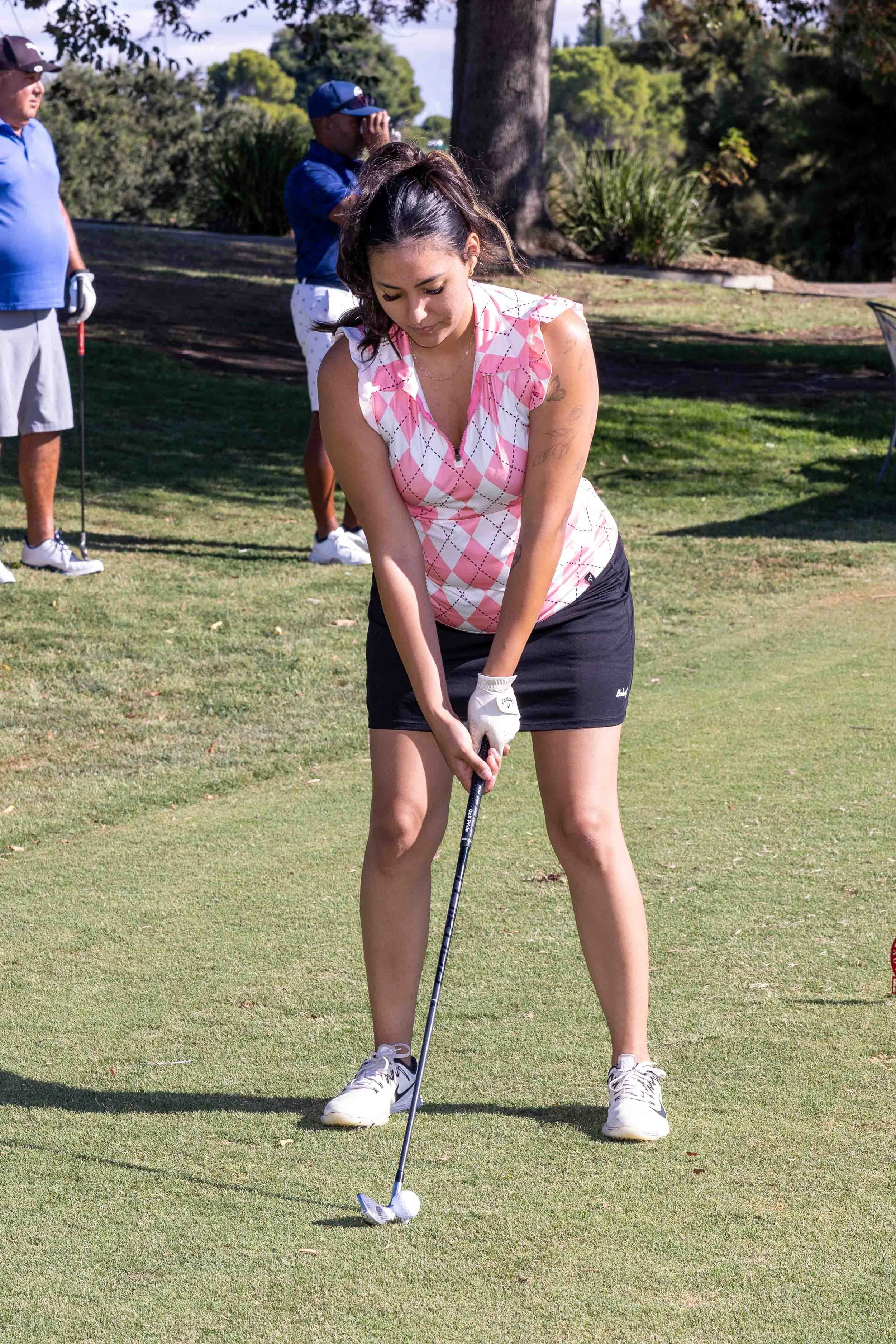 A woman with dark hair in a ponytail, wearing a pink and white argyle patterned sleeveless top, black shorts, and white golf shoes, preparing to swing a golf club on a lush green golf course with trees in the background. Two men are in the background