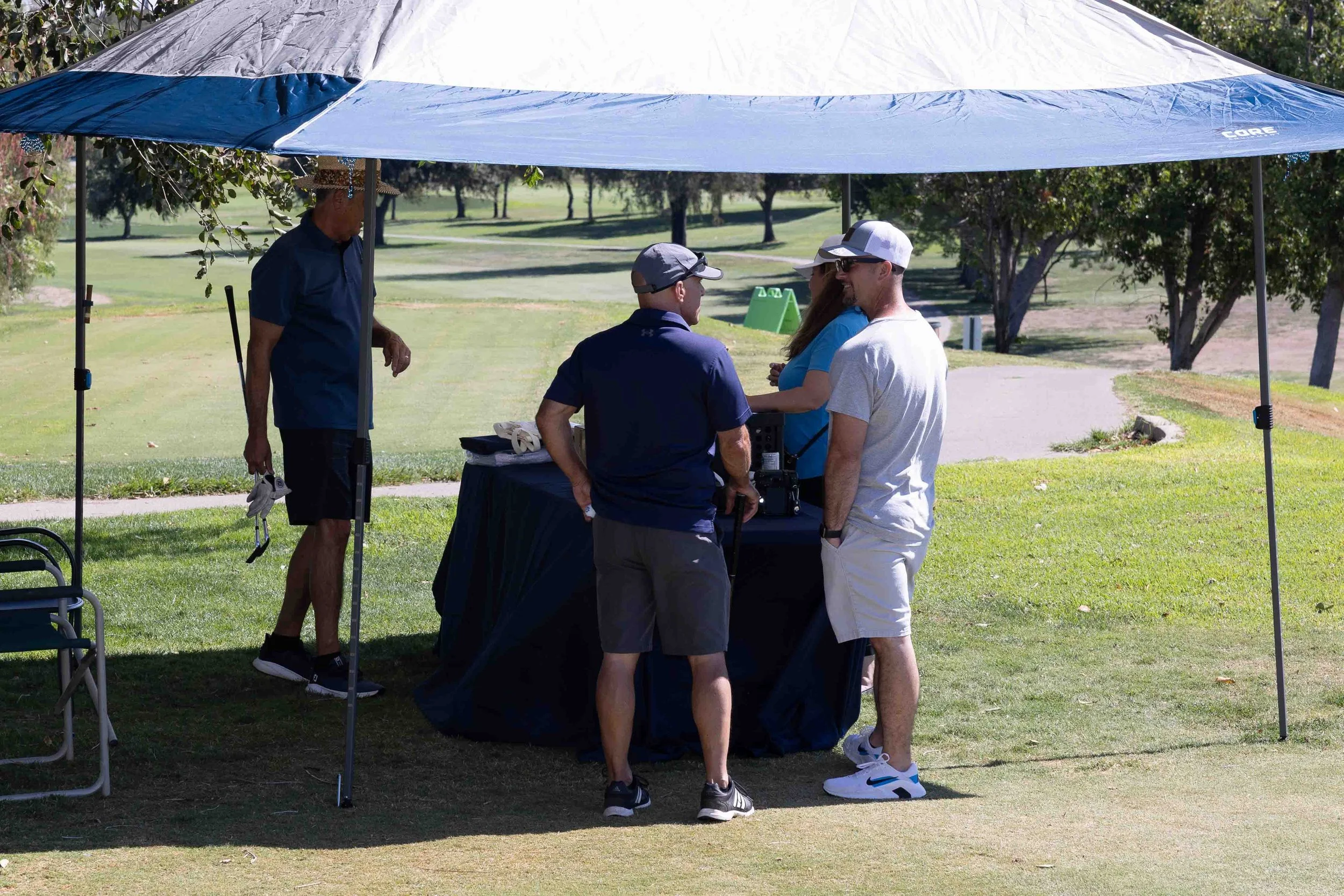 Four people under a large blue and gray umbrella outdoors, with a grassy park and trees in the background, engaging in conversation.