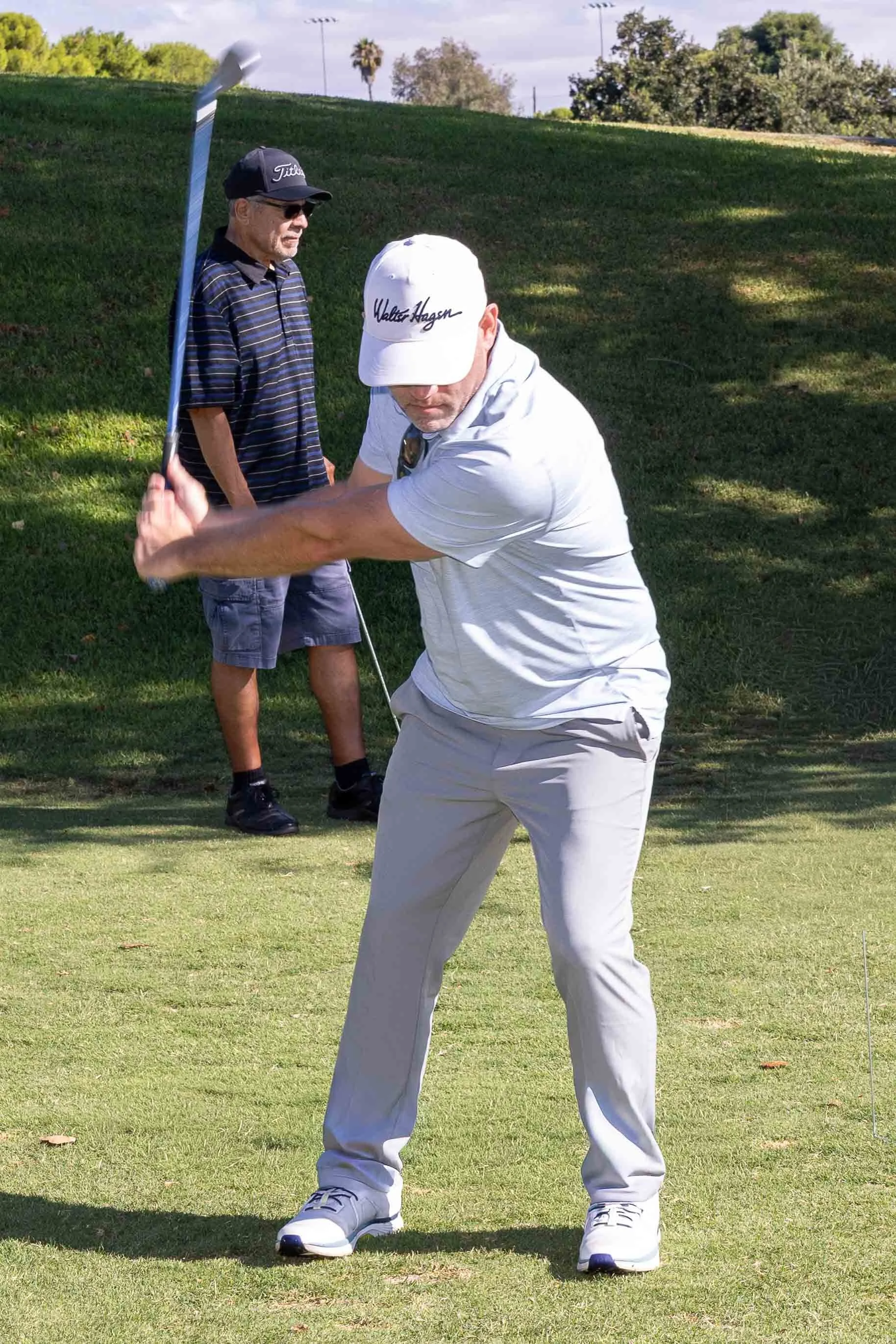 A man in a white cap and cream pants is swinging a golf club on a grassy golf course. Another man in sunglasses, a dark cap, and a striped shirt is standing behind him, watching. There are trees and light poles in the background, with partly cloudy s