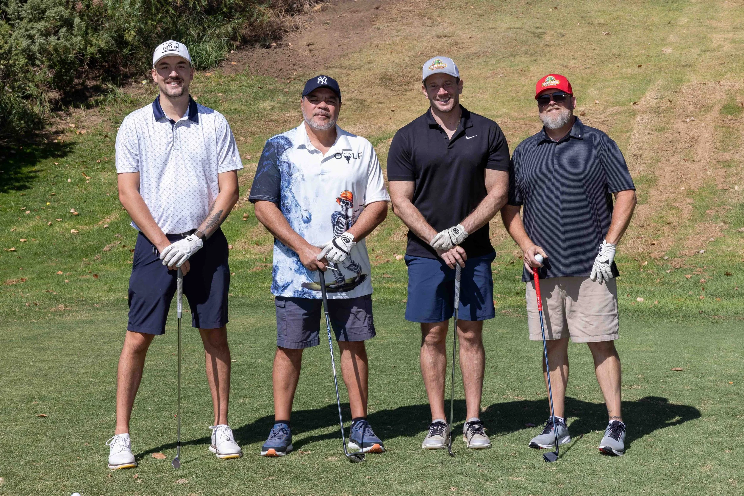 Four men standing on a golf course holding golf clubs, smiling, with greenery and a grassy hill in the background.