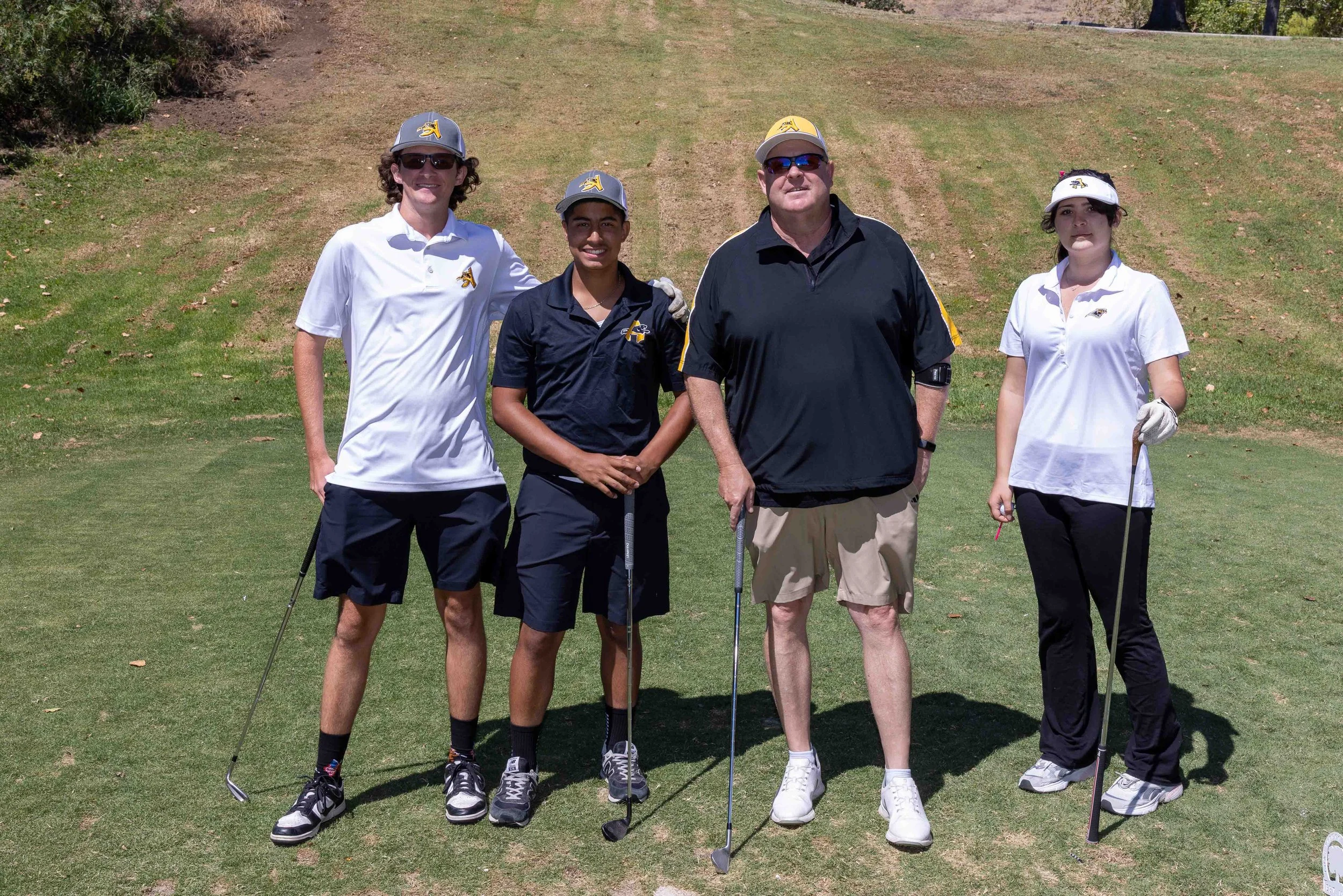 Four people standing on a golf course, holding golf clubs, wearing golf attire and hats, with a grassy hill in the background.