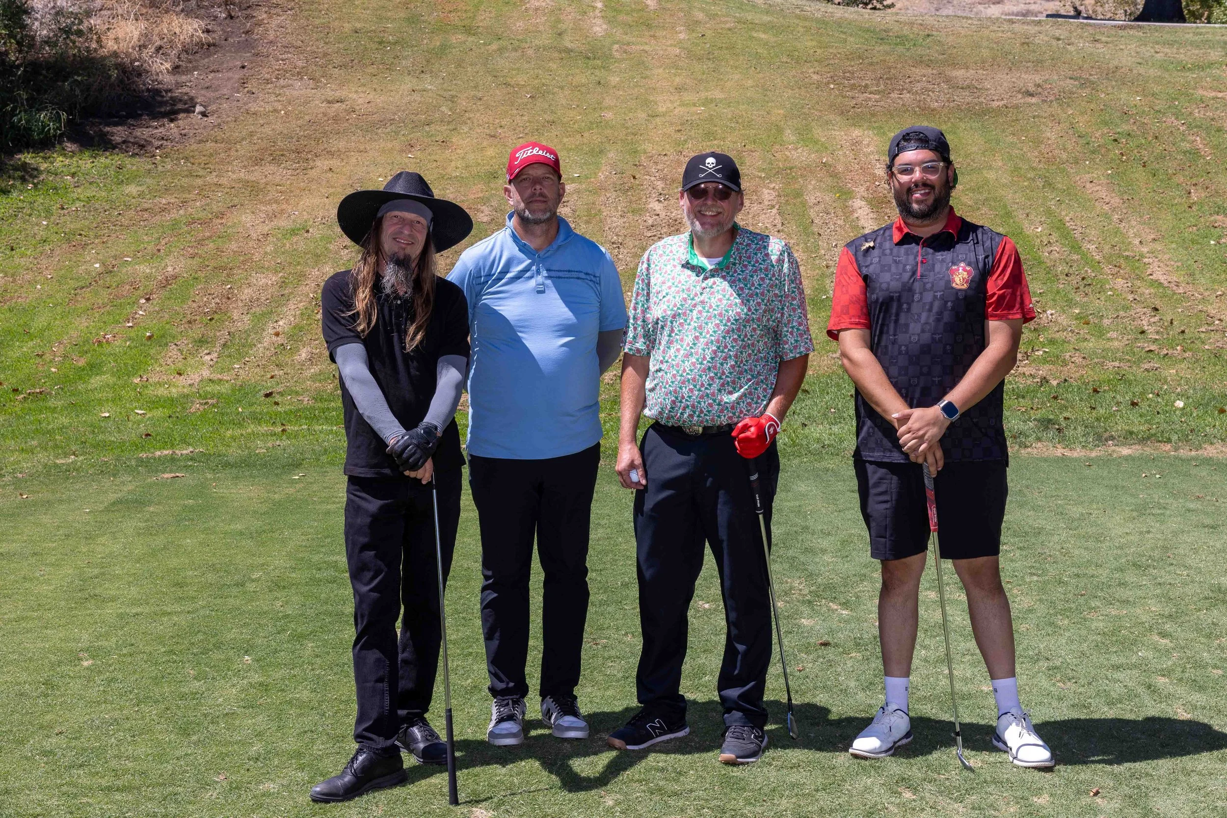 Four men standing on a golf course, holding golf clubs, dressed in casual golf attire, with green grass and a slightly sloped landscape in the background.