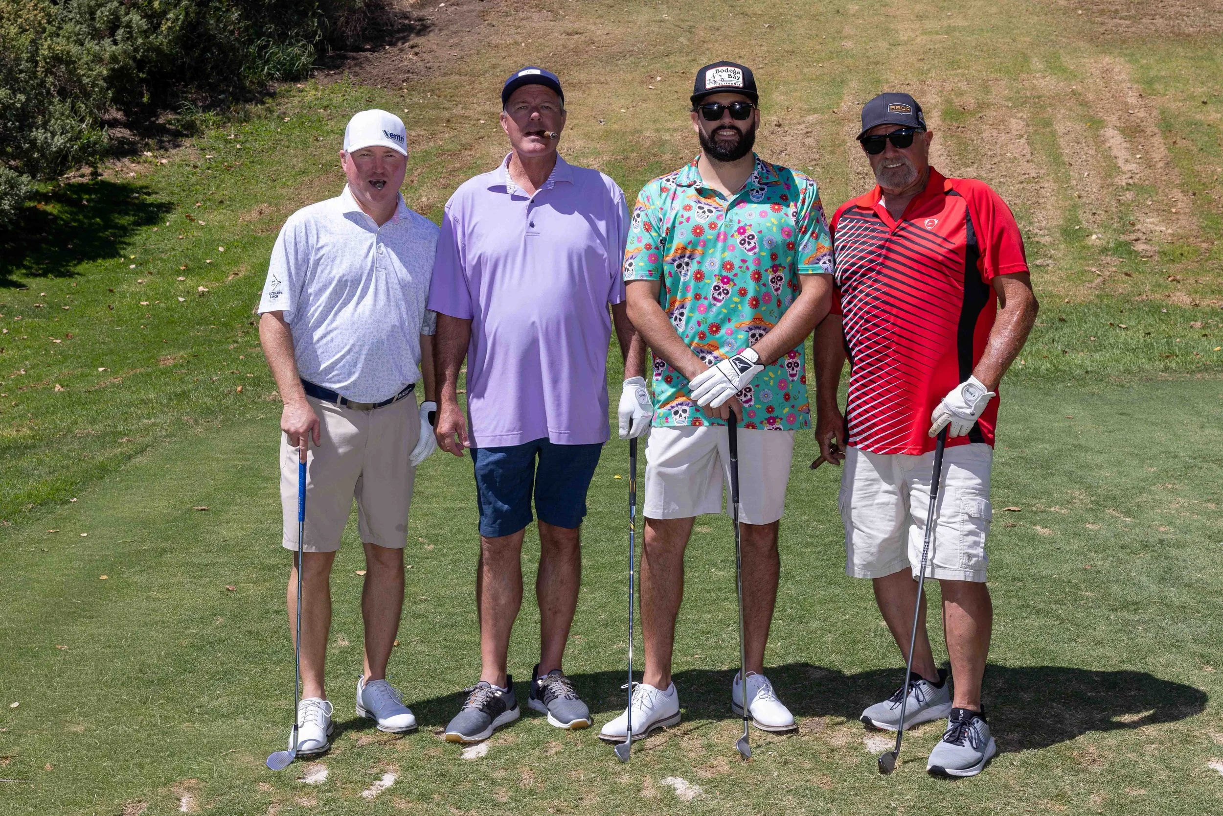 Four men standing on a golf course, holding golf clubs, dressed in casual golf attire, with trees and grass in the background, sunny day.