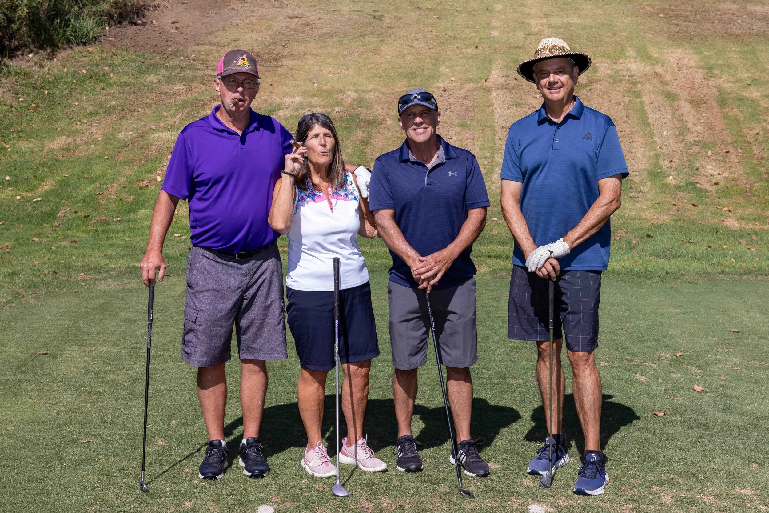 Four people standing on a golf course, holding golf clubs, dressed in athletic golf attire, with a grassy background.
