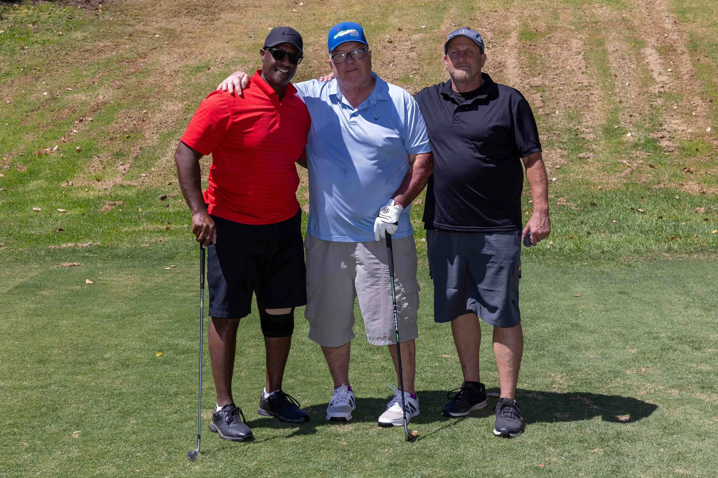 Three men standing together on a golf course, smiling, with golf clubs in hand, wearing athletic clothes and hats, under a clear sky.