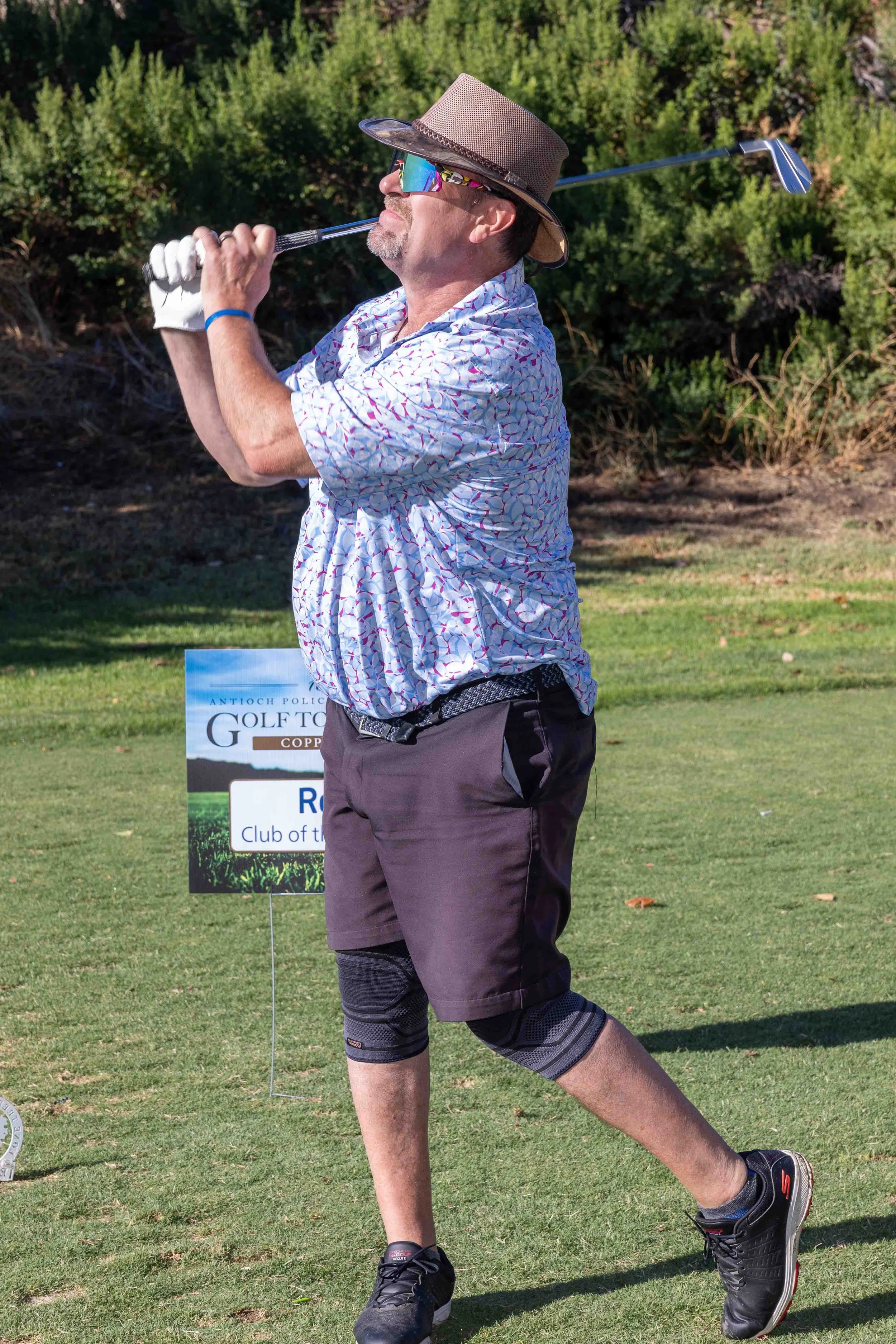 A man dressed in a floral patterned shirt, shorts, and athletic shoes, wearing a wide-brimmed hat, sunglasses, and a golf glove, is swinging a golf club outdoors on a sunny day. In the background, there is a sign for a golf tournament and a lush gree