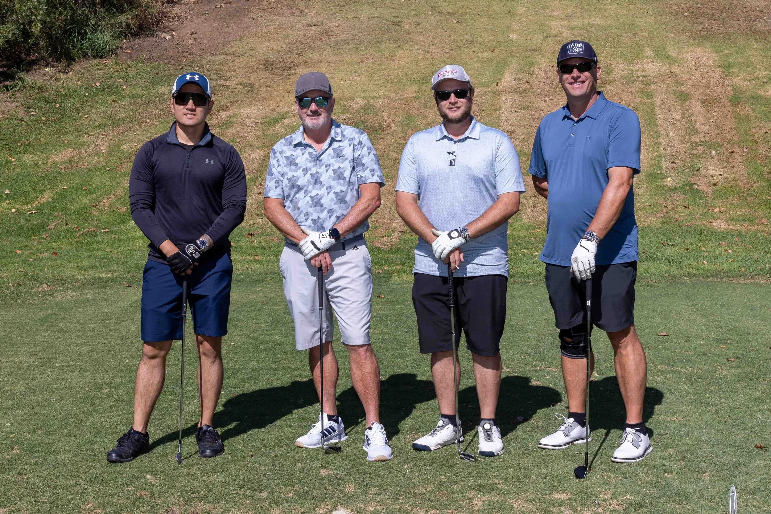 Four men standing on a golf course holding golf clubs, dressed in golf attire and sunglasses, with a grassy background.