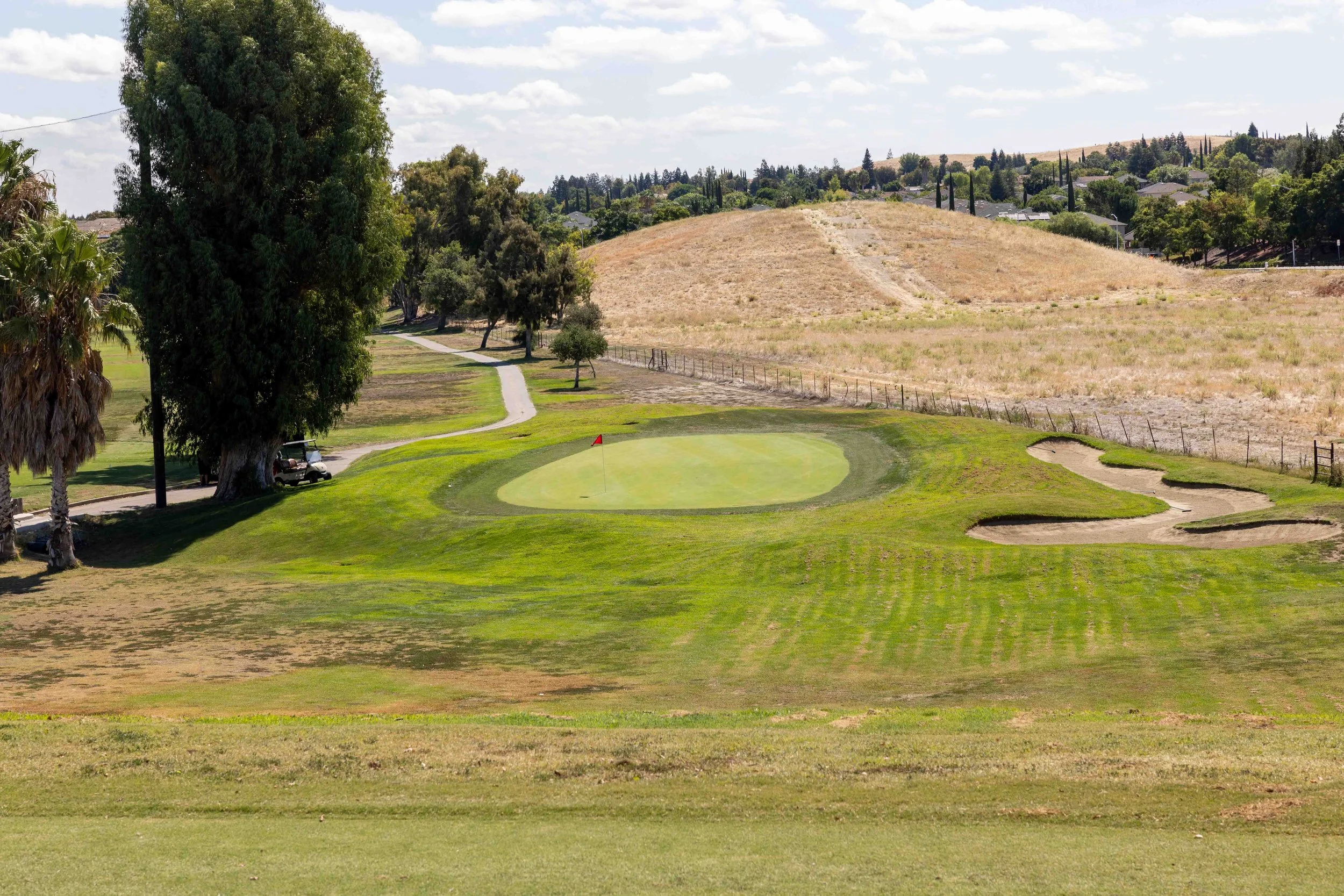 A golf course with a putting green, surrounded by trees and a trail, with hills and houses in the background under a partly cloudy sky.