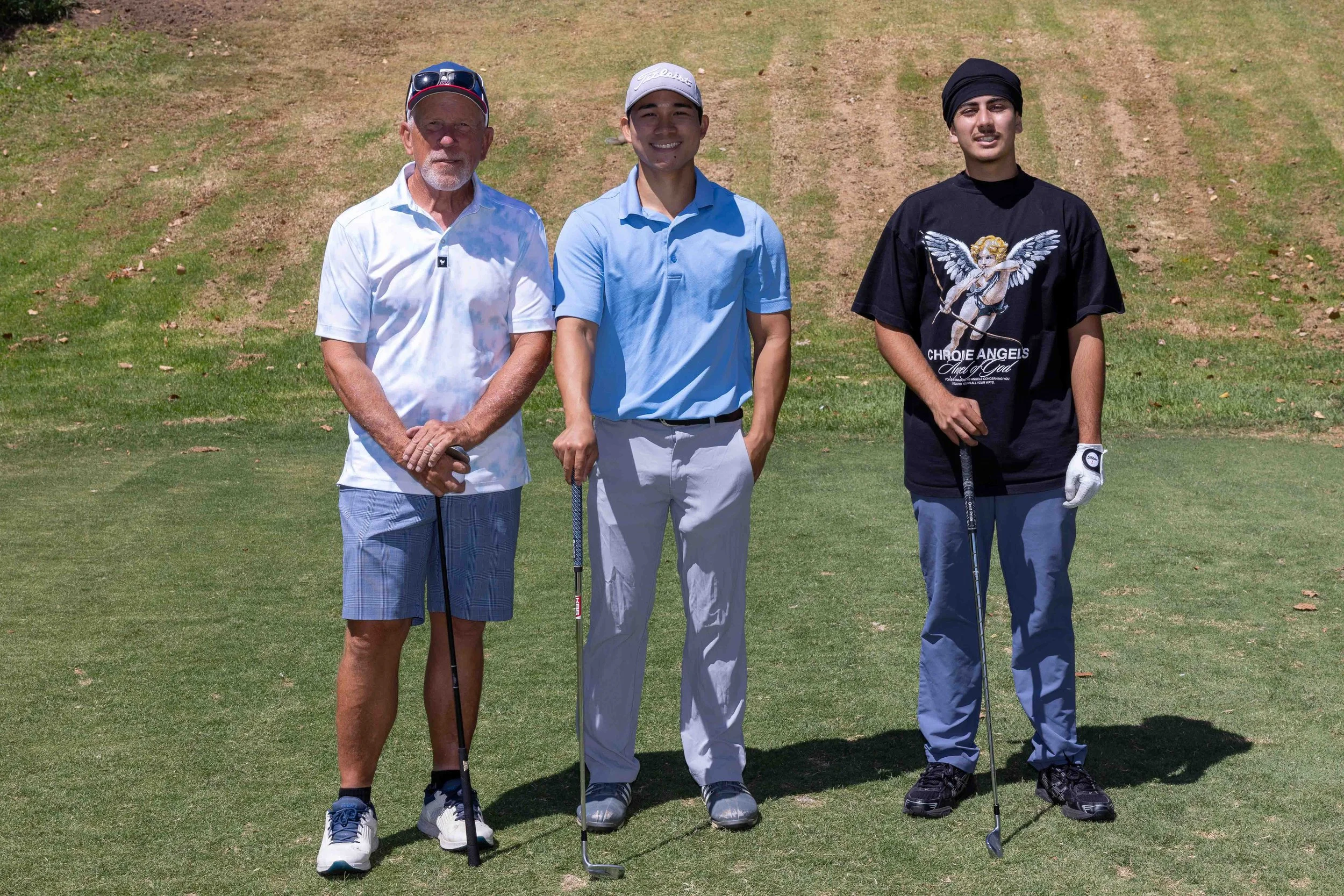Three men standing on a golf course holding golf clubs, smiling at the camera. The man on the left wears a white polo shirt, plaid shorts, and a cap with sunglasses on it. The man in the middle wears a light blue polo shirt and light-colored pants. T