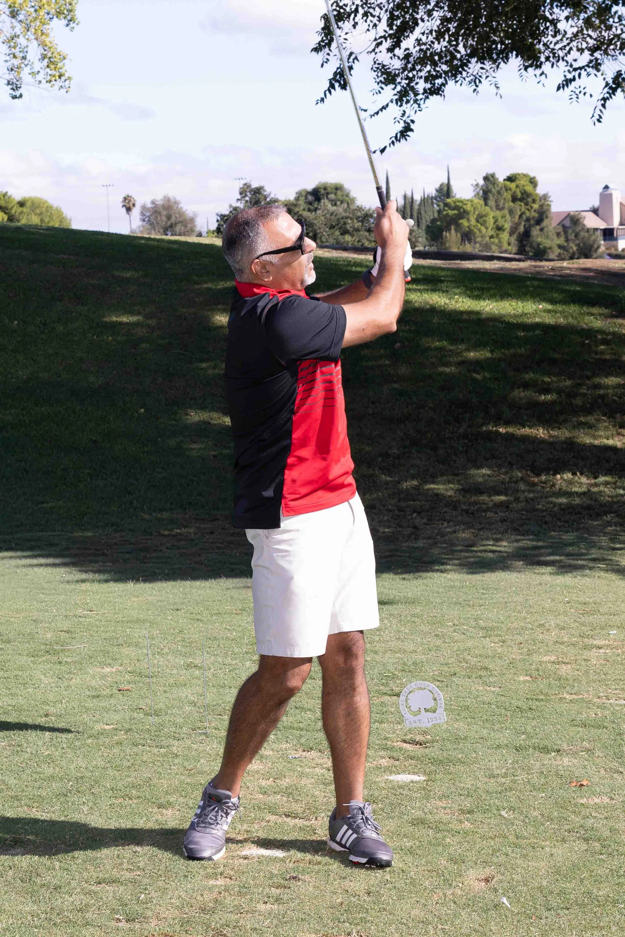 A middle-aged man with gray hair and sunglasses, wearing a black and red polo shirt, white shorts, and sports shoes, is preparing to tee off on a golf course. He is holding a golf club and standing on a well-maintained grassy area under a partly clou