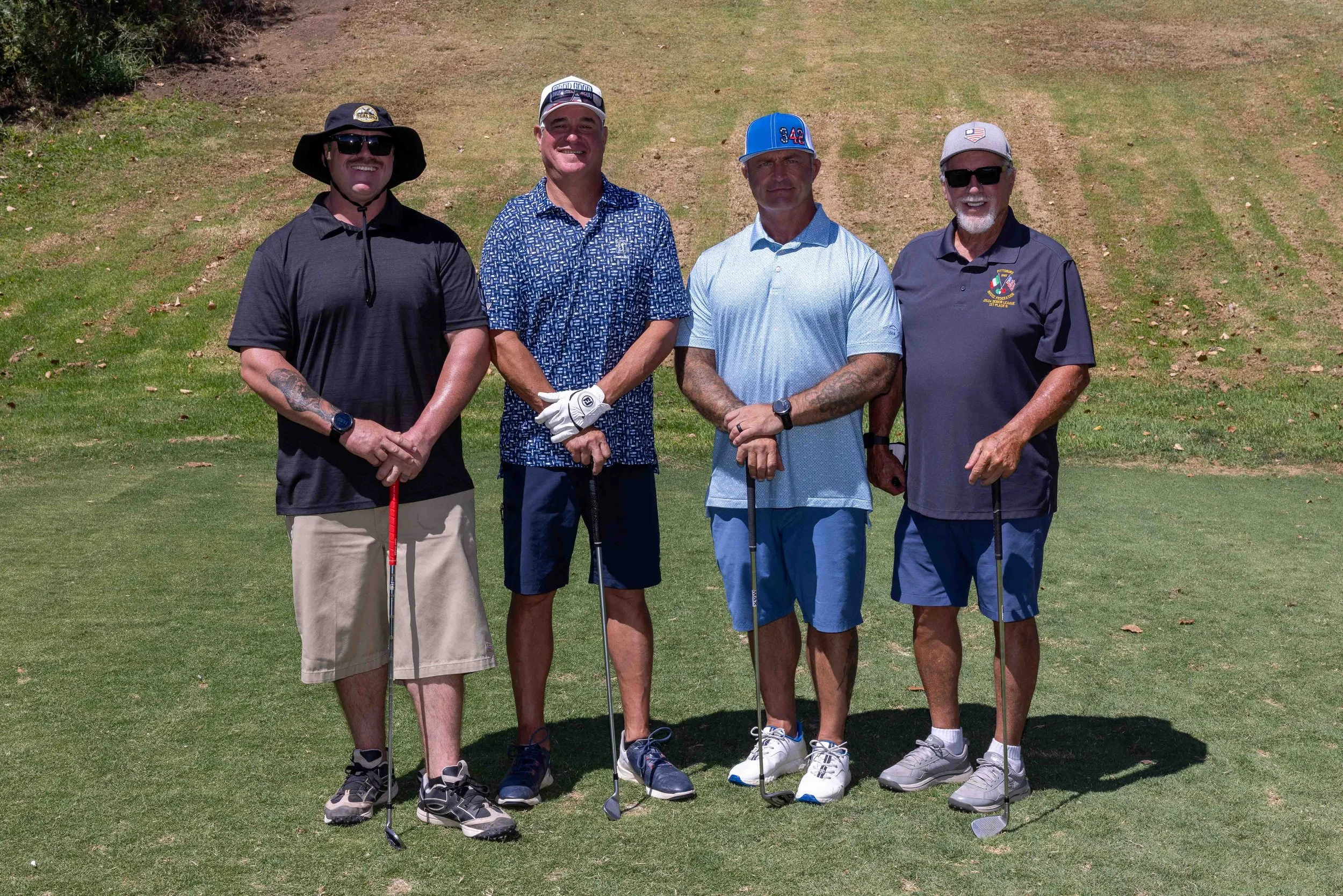 Four men standing on a golf course holding golf clubs, smiling at the camera during daytime, dressed in casual golf attire with hats and sunglasses.