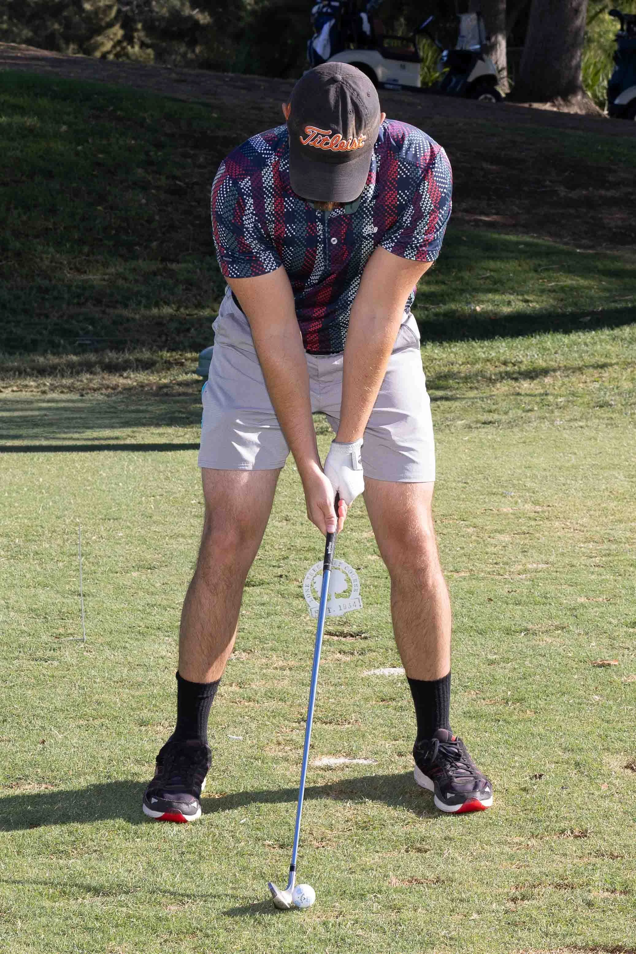A man wearing a gray titleist cap, a patterned tennis shirt, and shorts, standing on a golf course preparing to hit a golf ball with a club.