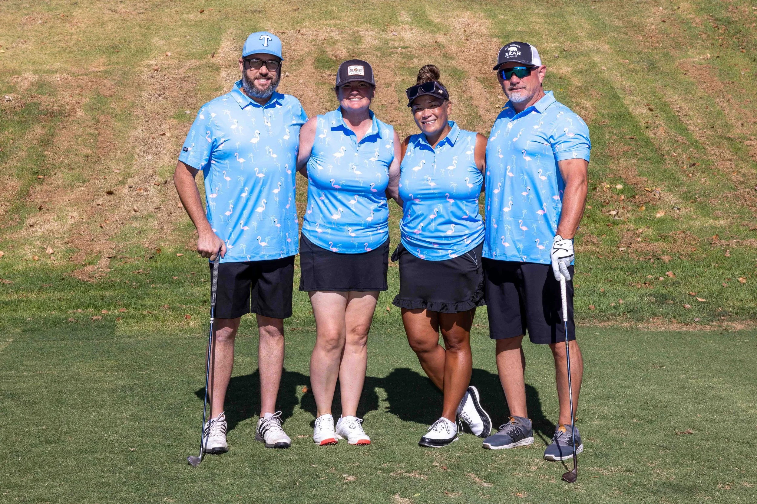 Four people standing on a golf course, dressed in matching blue flamingo shirts, smiling at the camera, with two men and two women, holding golf clubs.