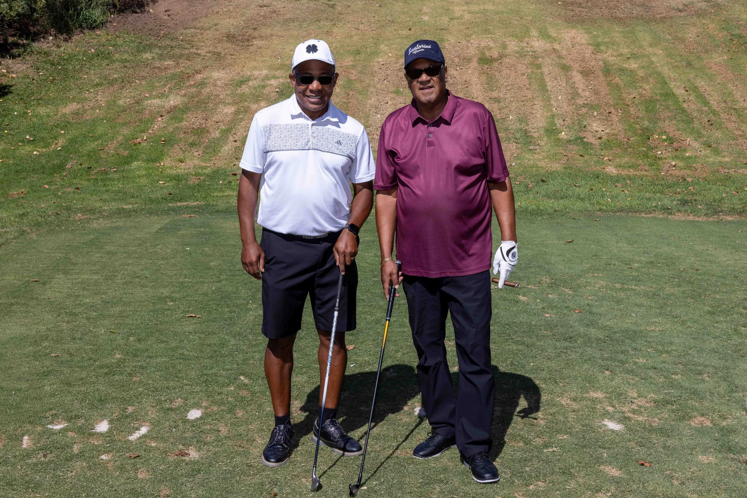 Two men standing on a golf course, holding golf clubs, dressed in golf attire with hats and sunglasses.