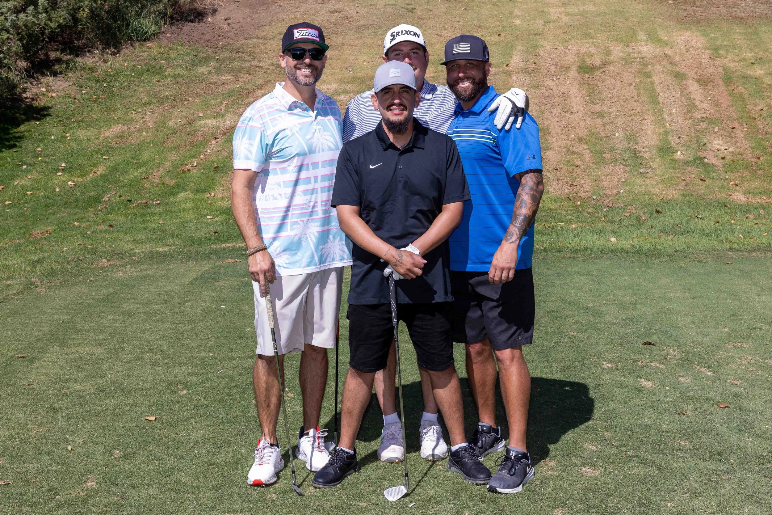 Four men standing on a golf course, smiling at the camera, holding golf clubs. They are wearing casual golf attire, including hats, sunglasses, and gloves. The background features grass and a small hill.