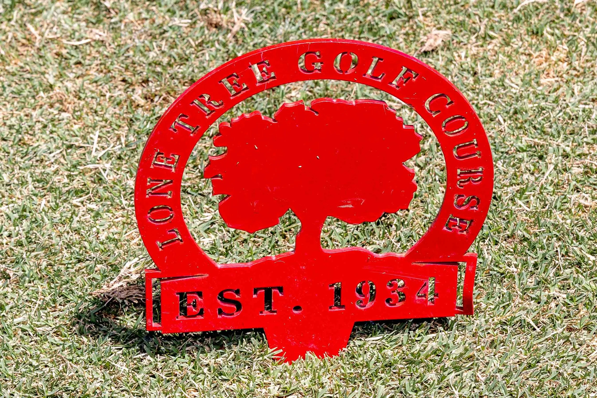 Red metal sign with a tree logo and text indicating 'LONE TREE GOLF COURSE EST. 1934' on a grassy surface.