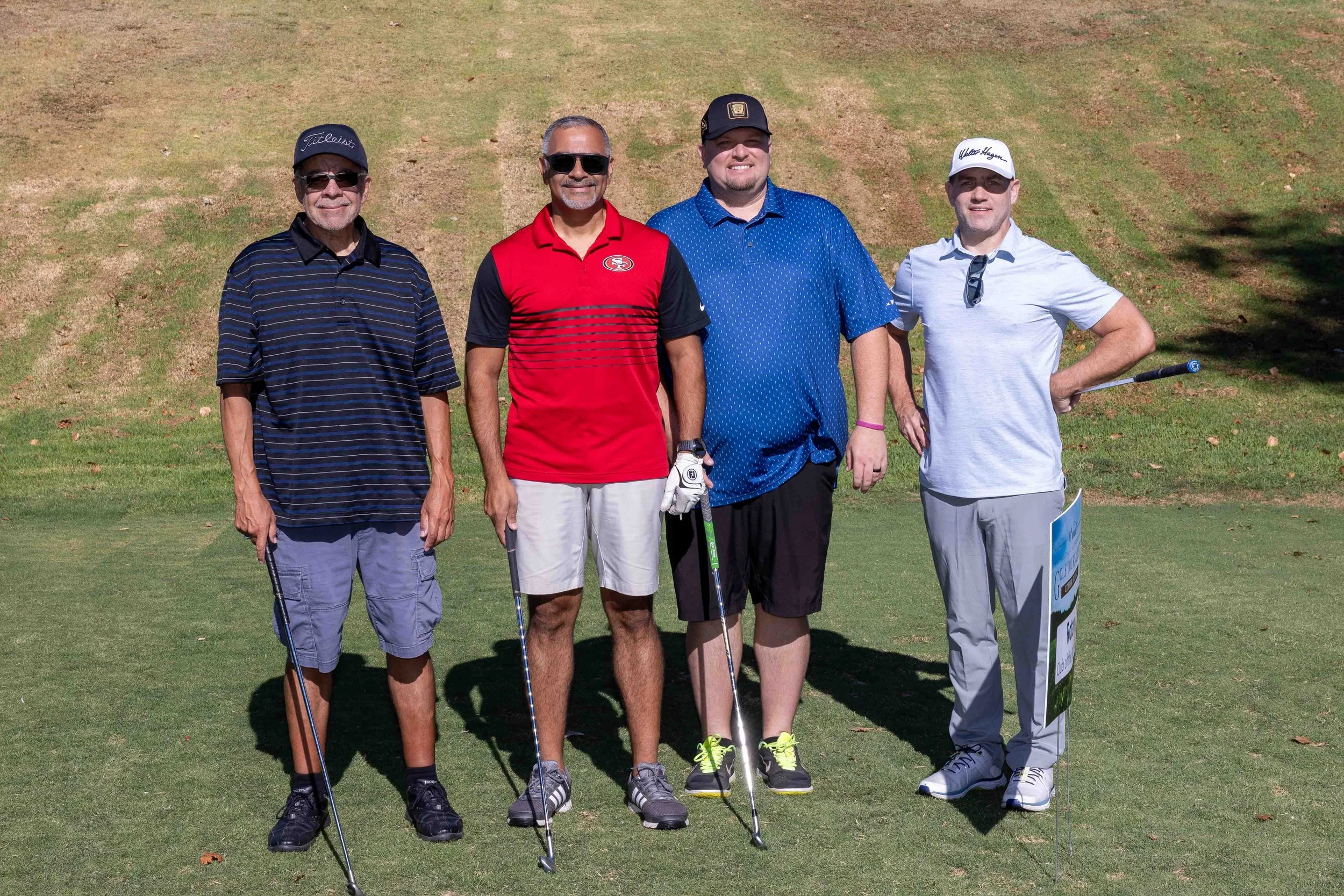 Four men standing on a golf course holding golf clubs, wearing casual golf attire and sunglasses.