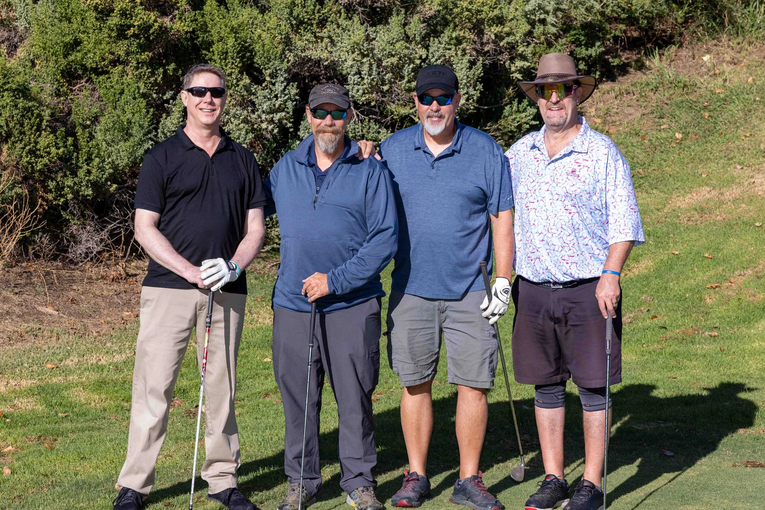 Four men standing on a golf course with trees and grass in the background, holding golf clubs.