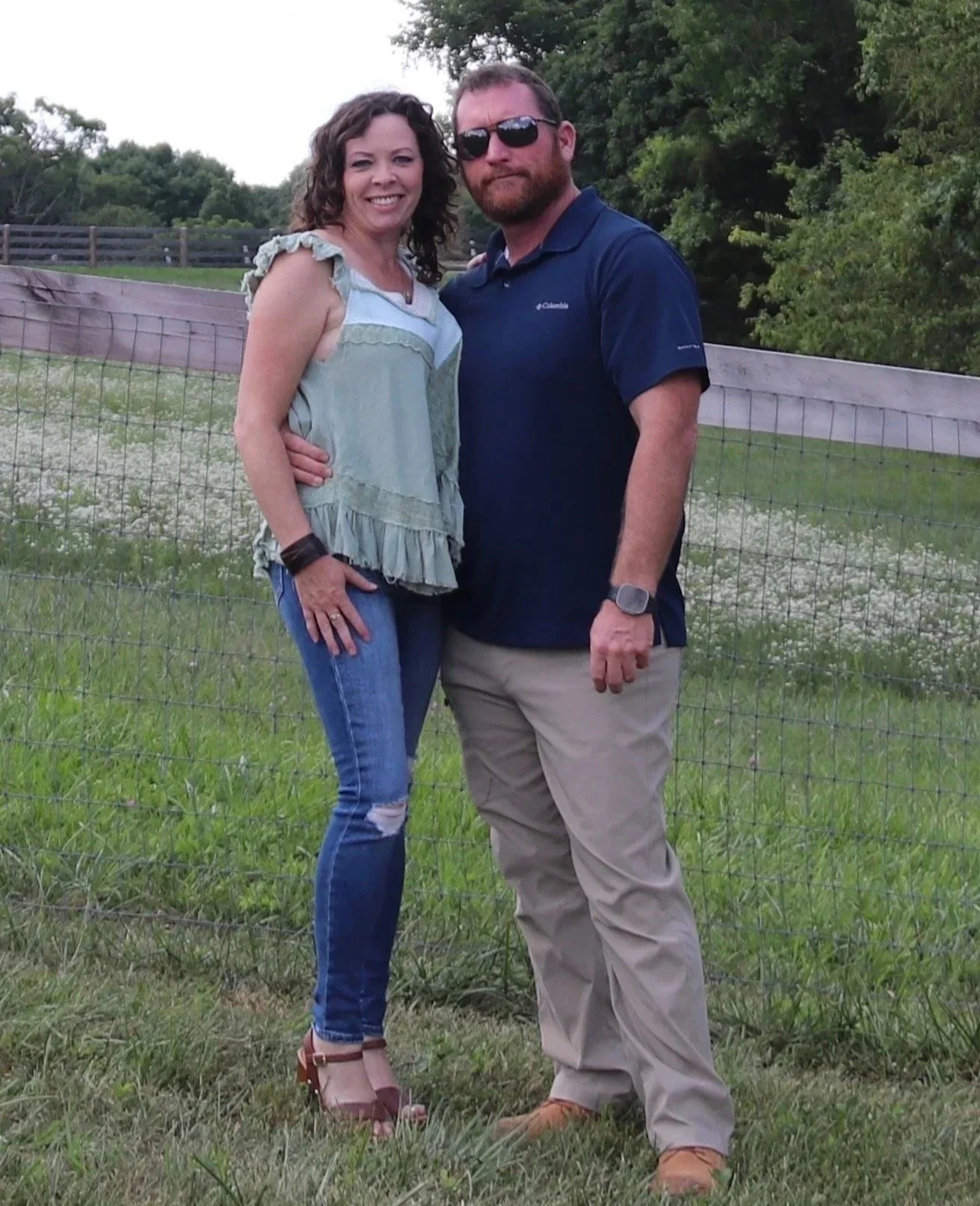 A smiling woman and a man in sunglasses standing outdoors near a fence, with trees and grassy field in the background.