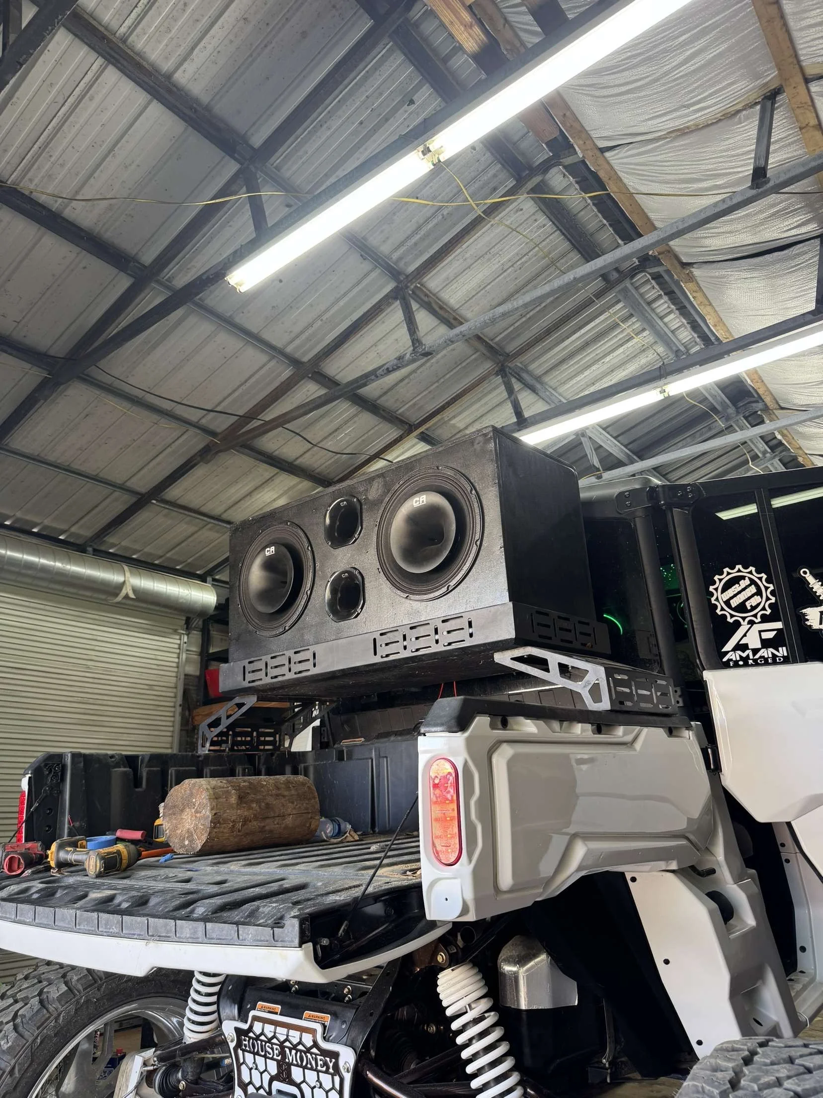 Large black speaker with two woofers and two tweeters mounted on the back of a utility vehicle in a garage or workshop.