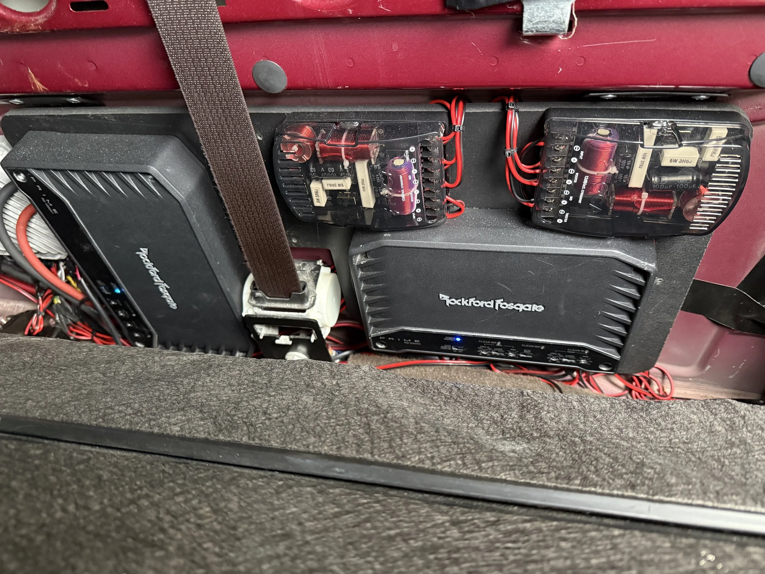 Electrical setup with two black Rockford Fosgate power amplifiers, two clear plastic tube fuses with red wiring, and a black amplifier with a blue LED indicator, all secured on a red surface with black straps.