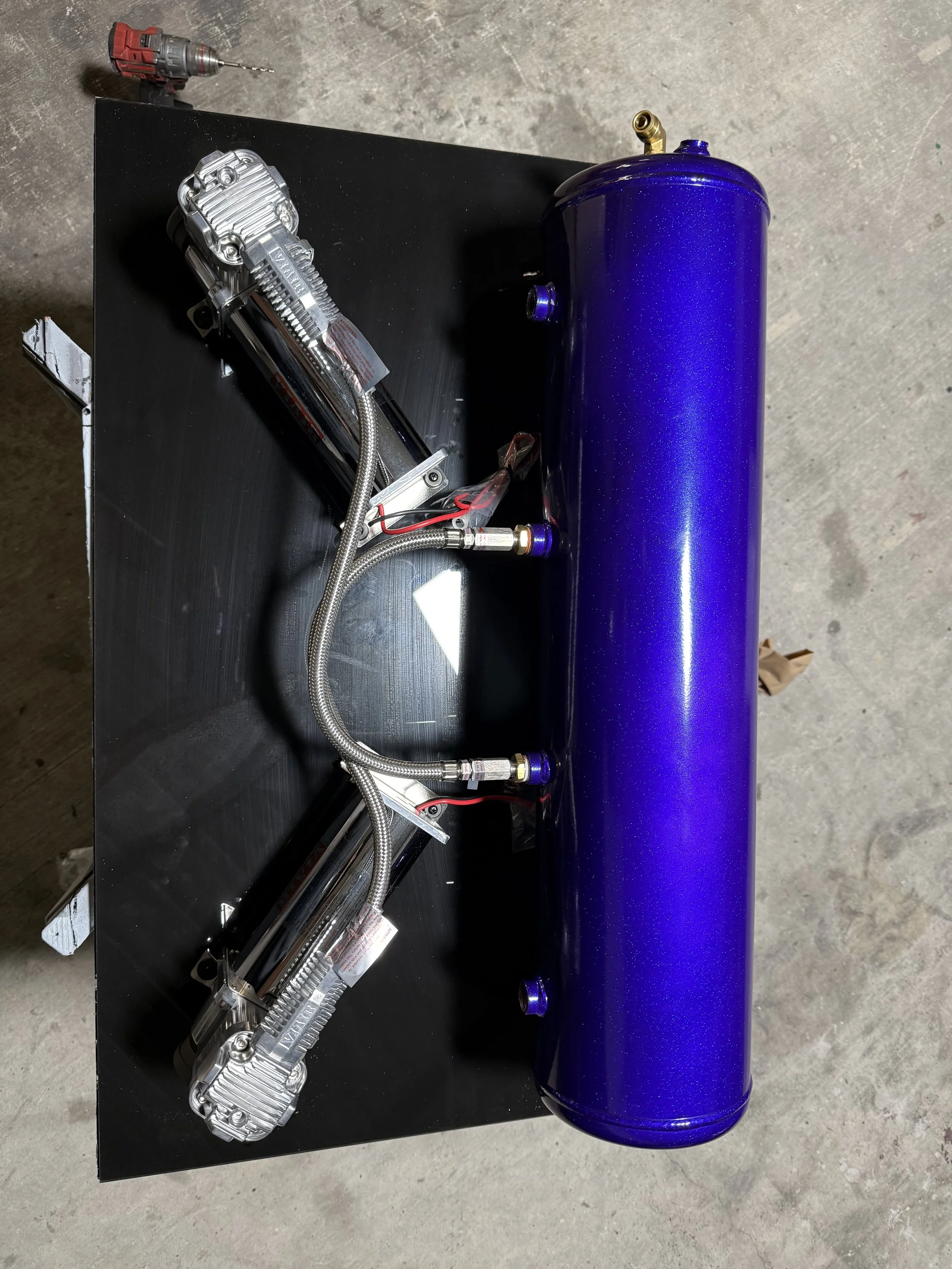 A blue anodized component connected to two silver turbochargers with braided lines, mounted on a black metal surface.