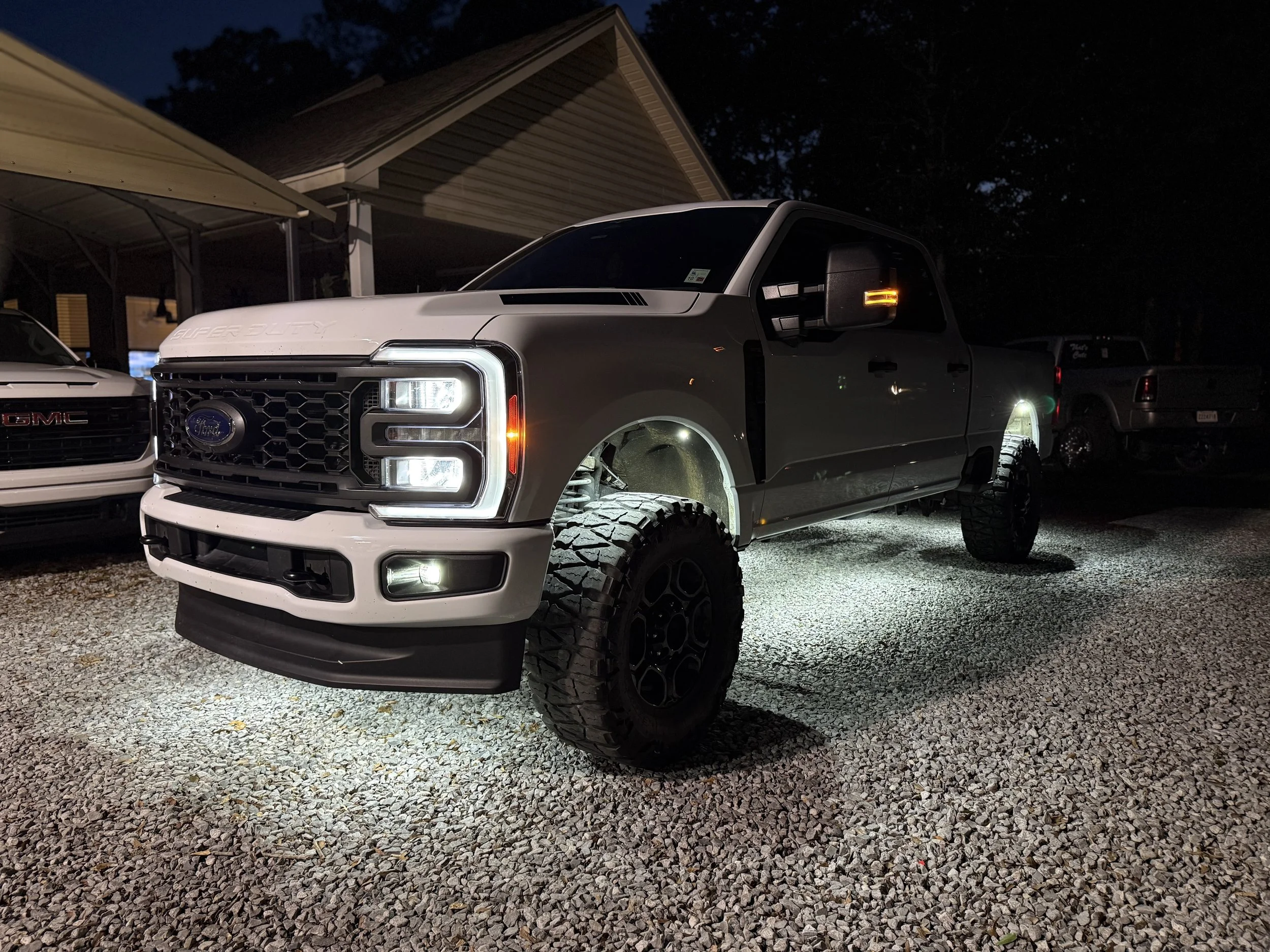 White Ford Super Duty truck with lifted suspension and large off-road tires parked on gravel at night, illuminated by its lights.