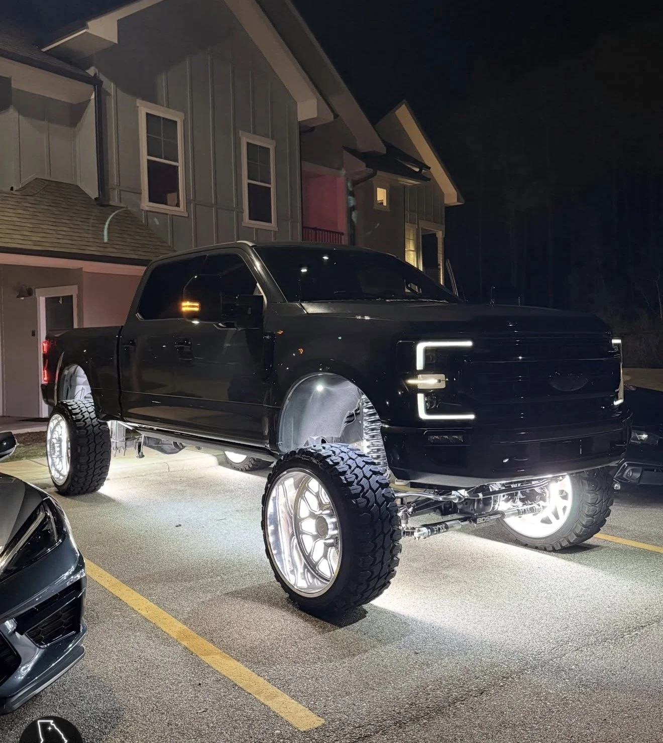 A black lifted pickup truck with large off-road tires is parked in a parking lot at night. The truck has bright LED lights outlining parts of the vehicle, including the front grille and wheel wells. There are other cars visible in the nearby parking 