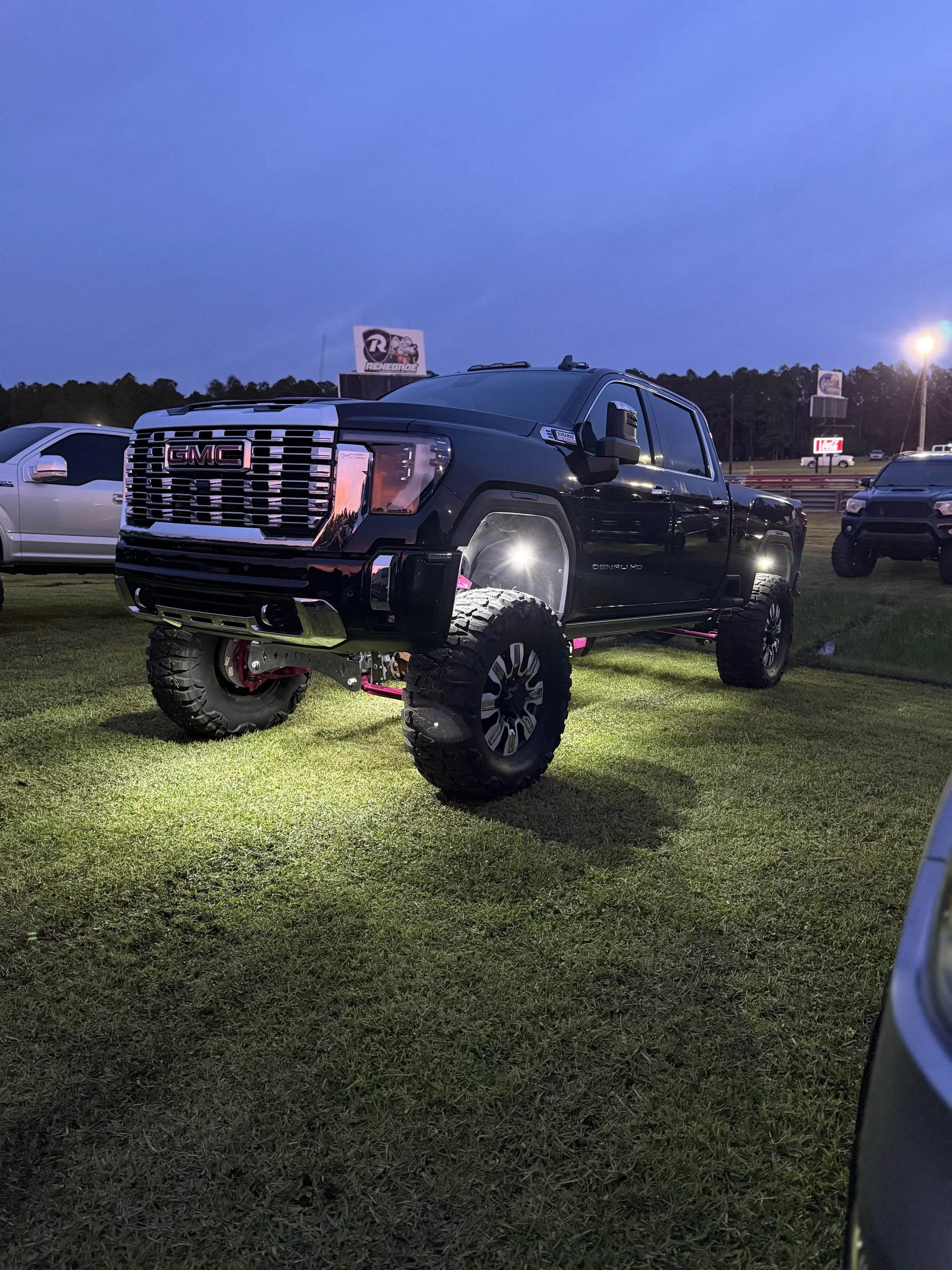 A black lifted GMC truck with off-road tires parked on grass at dusk, with other parked vehicles and a billboard in the background.