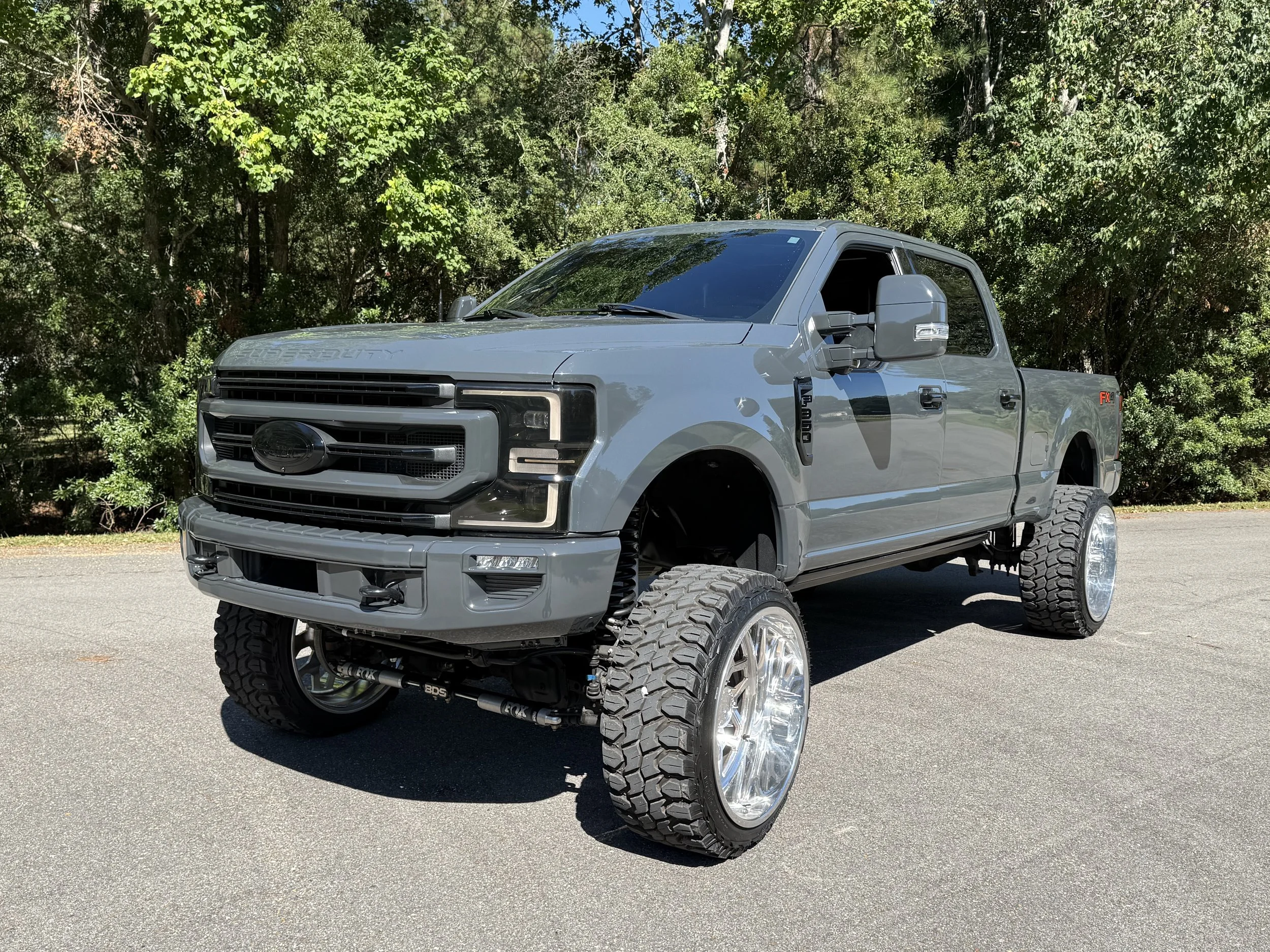 A gray lifted Ford F-250 truck with large off-road tires parked on pavement near green trees.
