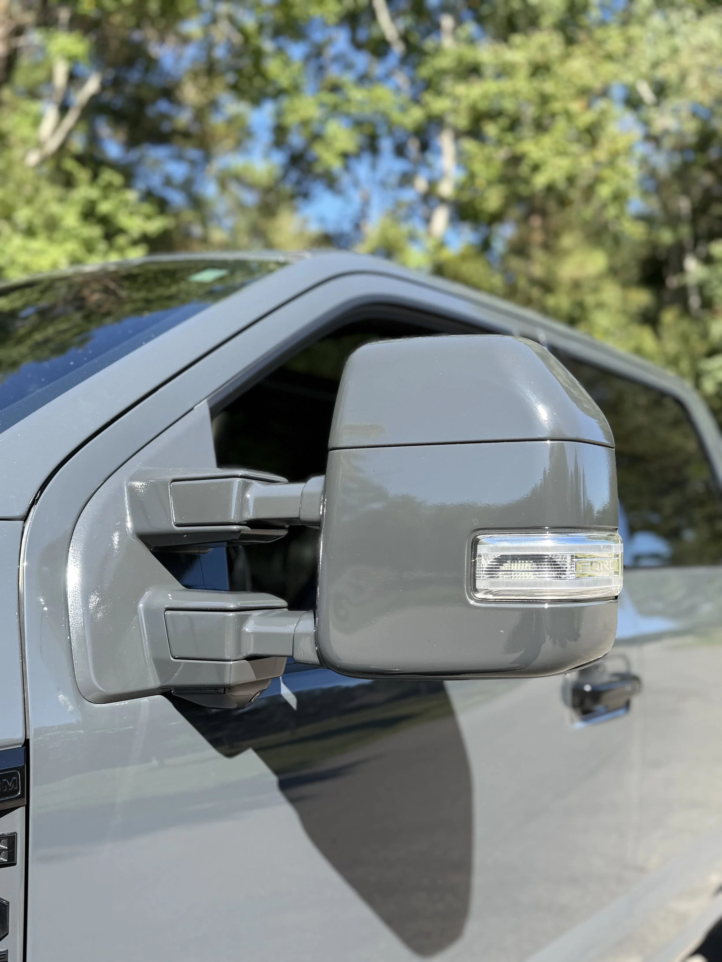 Close-up of a gray truck side mirror with trees and blue sky in the background.