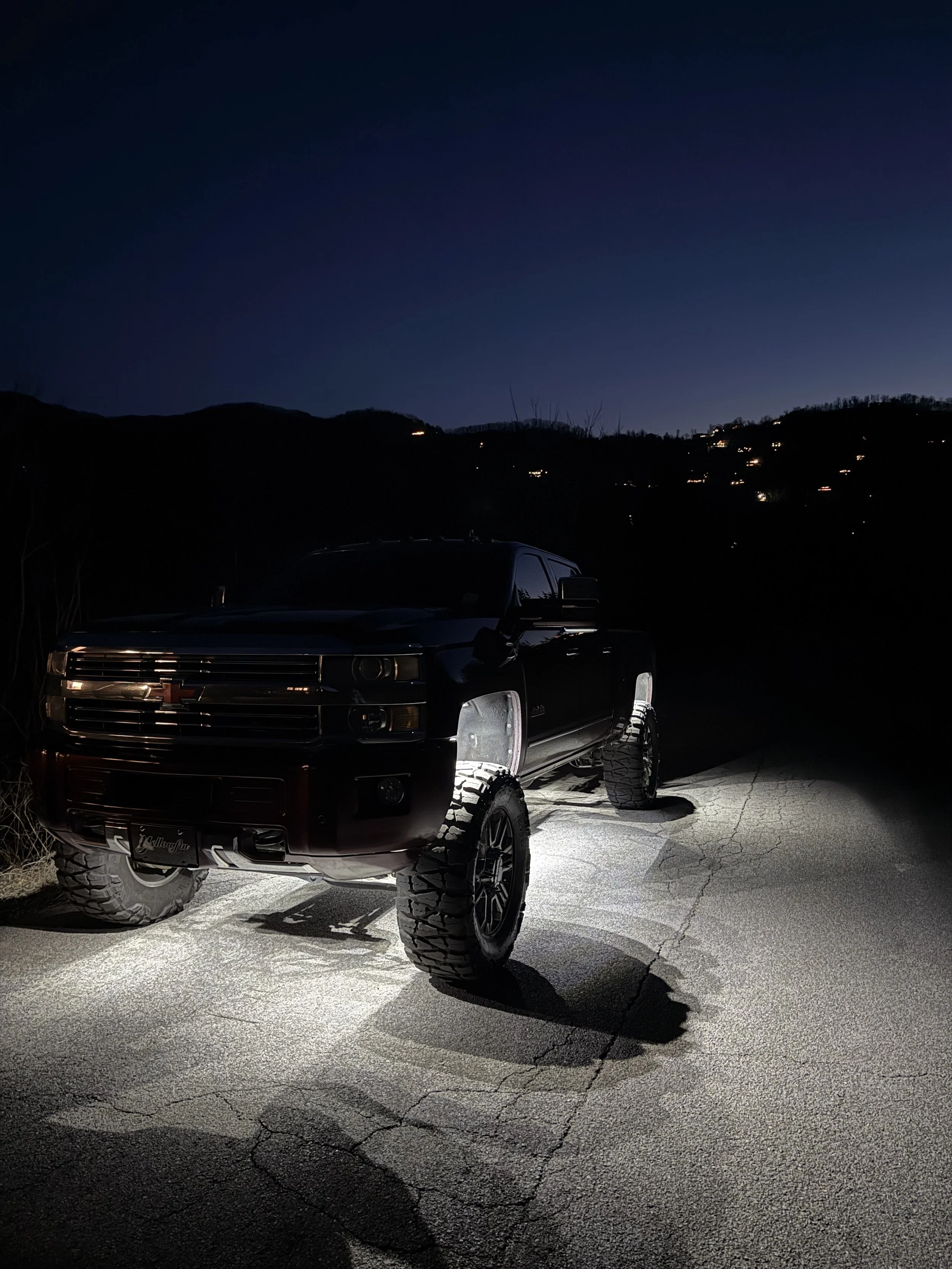 A black lifted pickup truck parked on a cracked road at night, with hills and scattered lights in the background, and an undefined sky above.