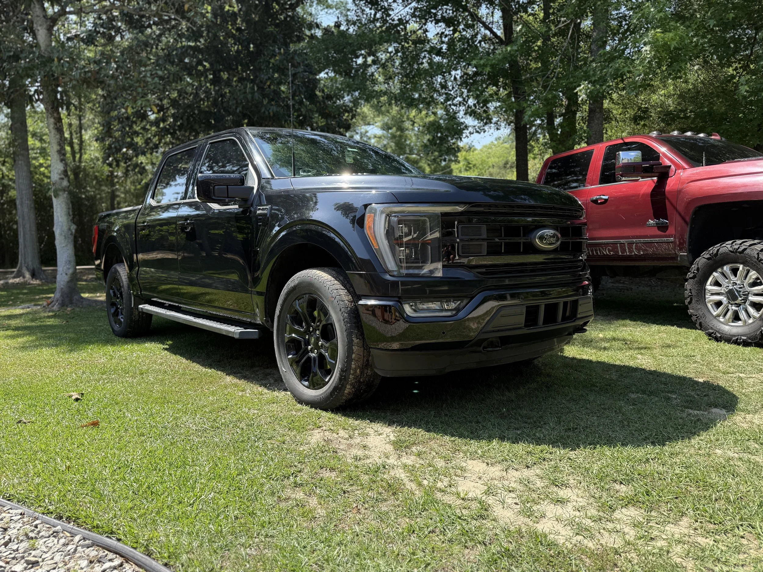 A black Ford pickup truck parked on grassy ground next to a red pickup truck in a wooded area.