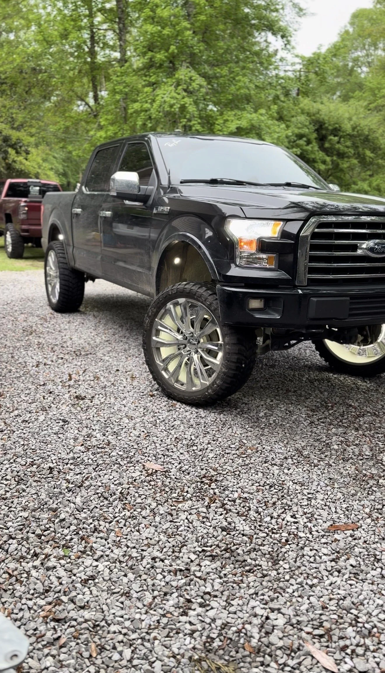 Black Ford F-150 truck with custom wheel rims parked on gravel surface, green trees in background.