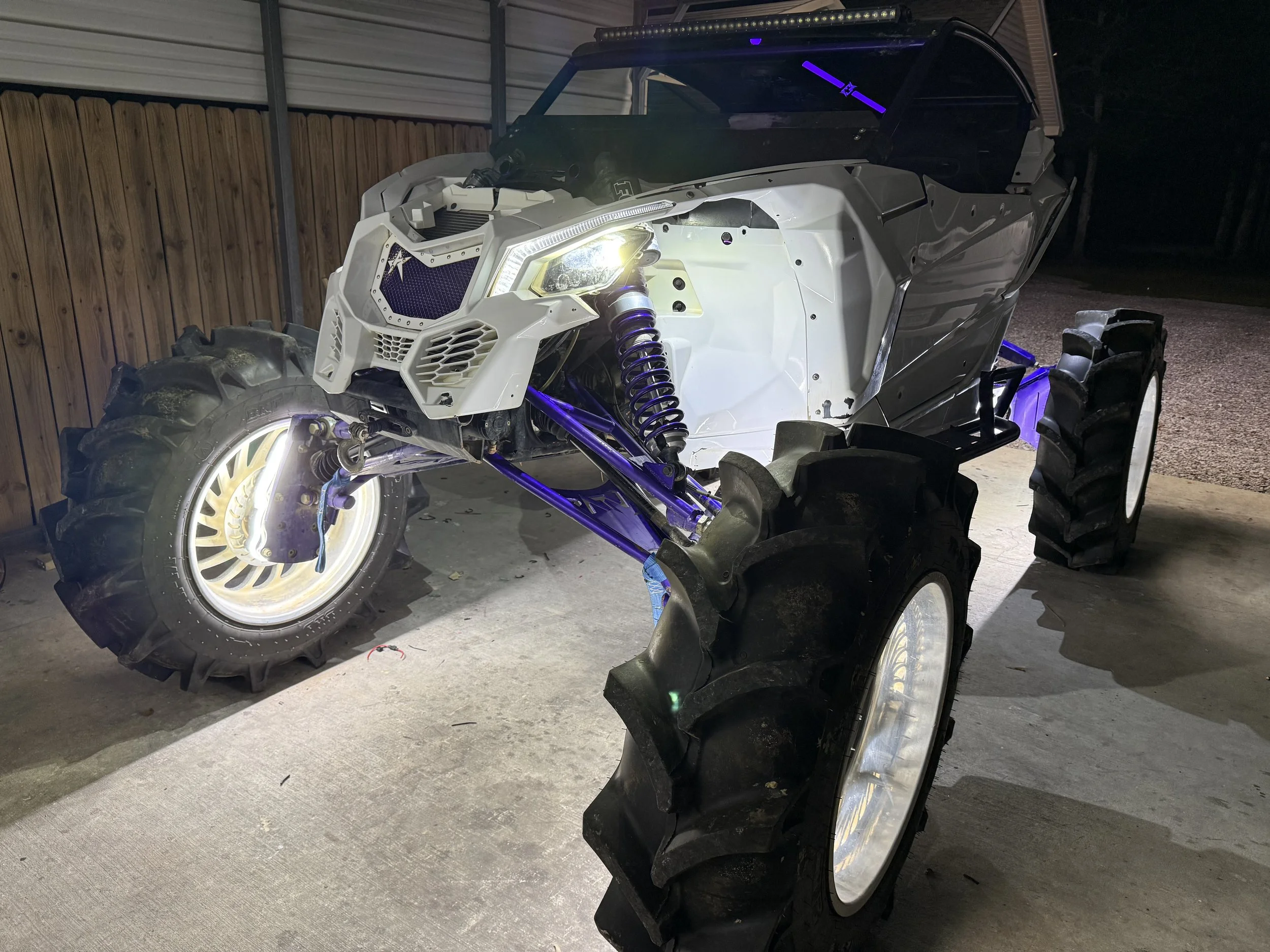 An off-road utility vehicle with large tires, a white body, and purple suspension parts is parked inside a garage with wooden and metal walls at night, illuminated by bright lights.