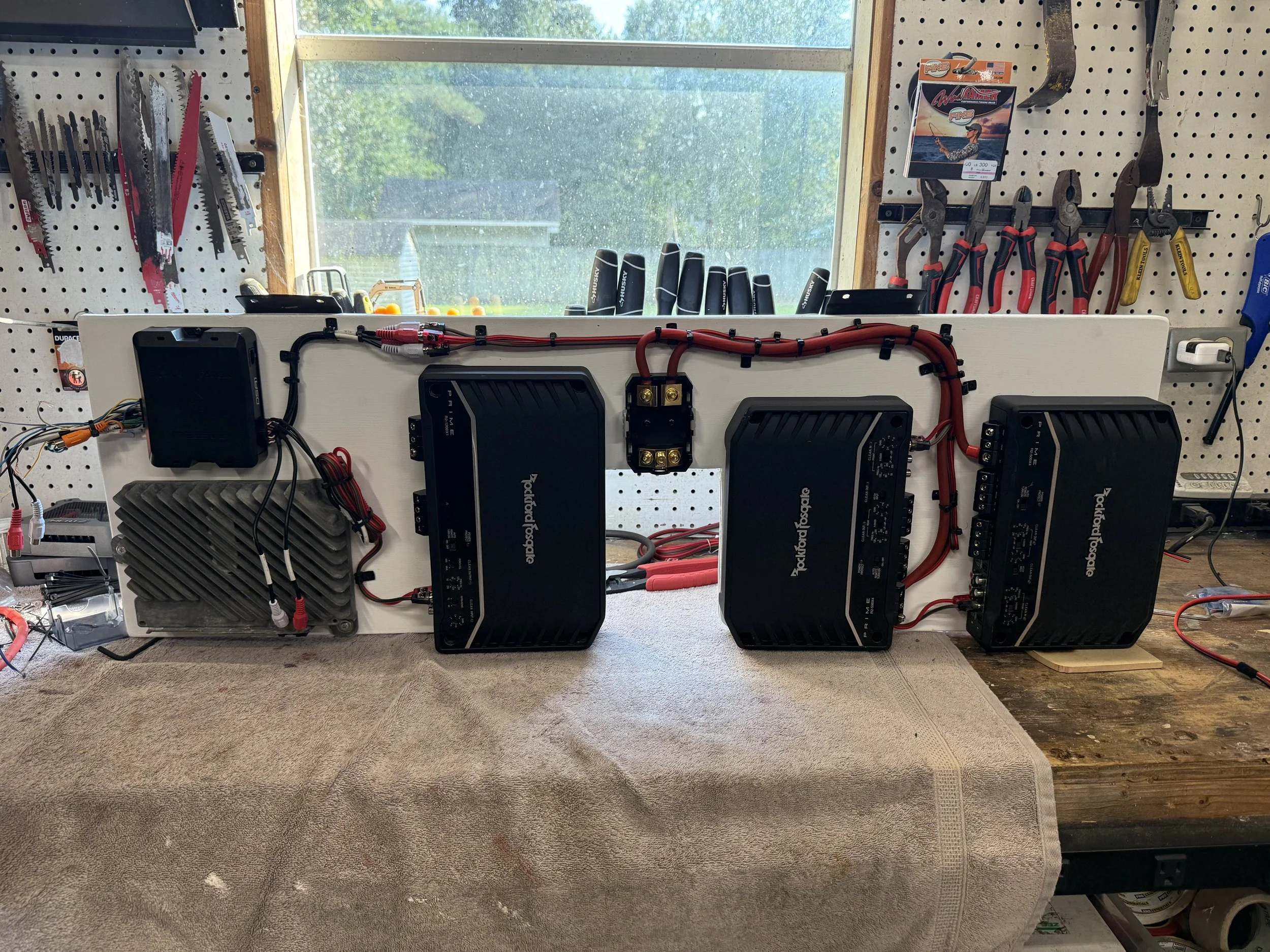 Electrical components and wiring on a white panel, mounted on a workbench in a workshop with tools hanging on a pegboard in the background.