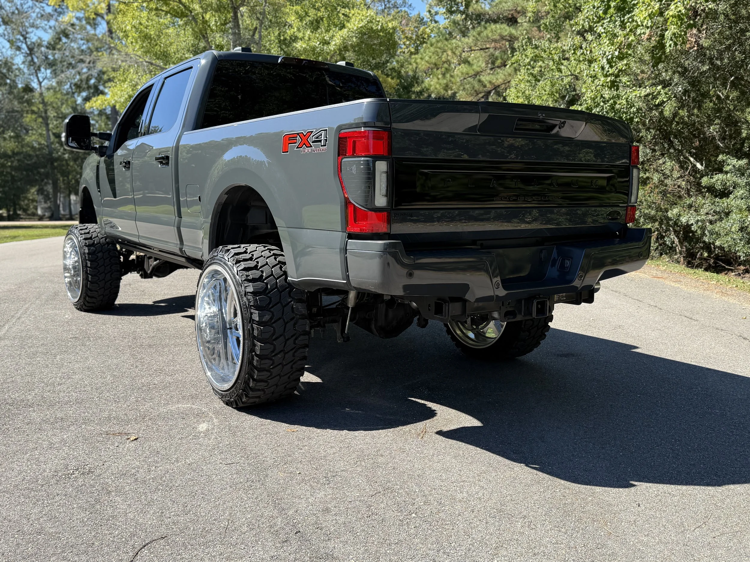 Gray pickup truck with oversized tires and chrome rims parked on asphalt, surrounded by green trees.
