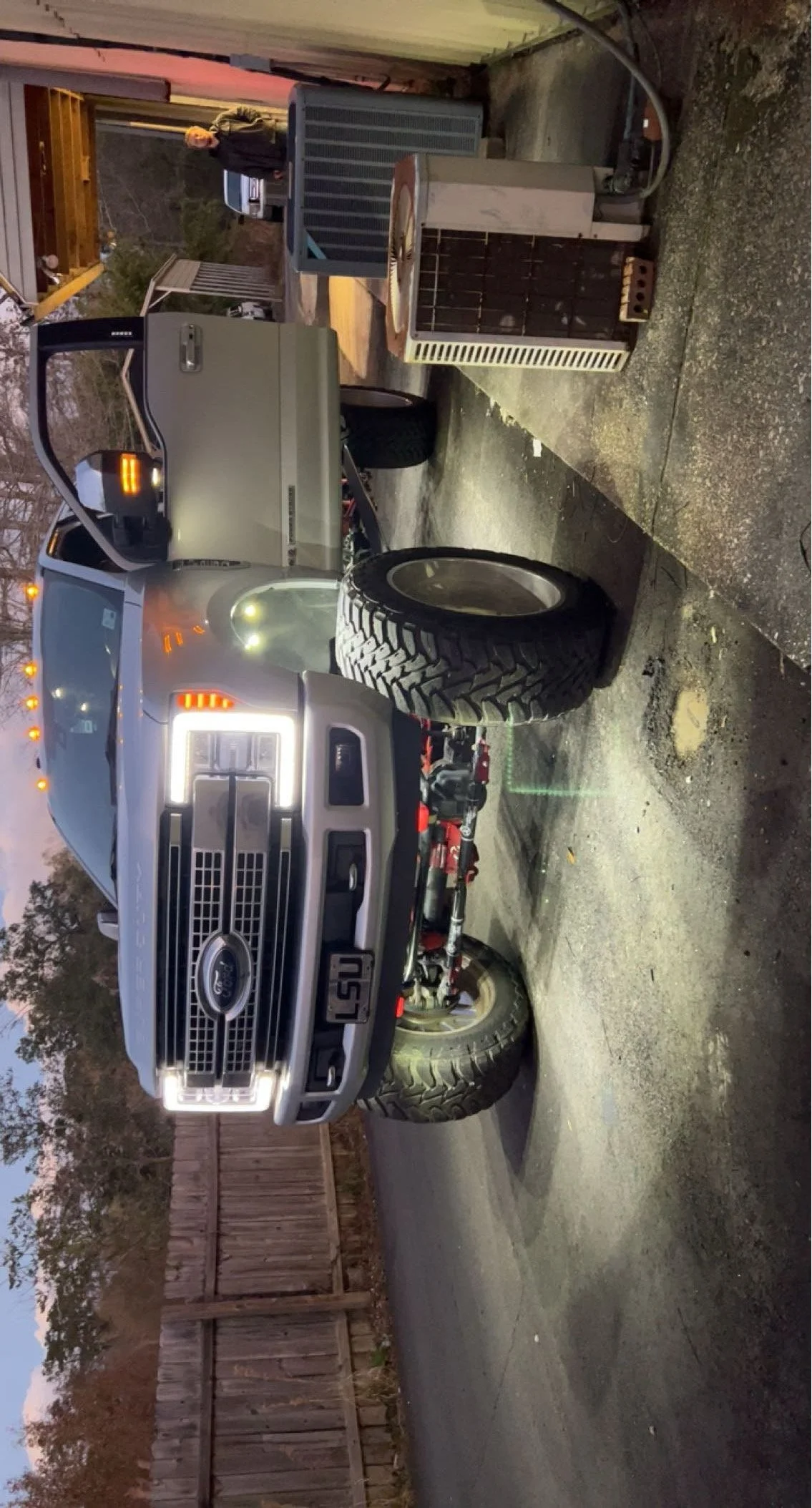 A black Ford truck with its front grille removed, revealing the engine, parked in a driveway. There are various tools and equipment nearby, including a heater and a person standing in the background.