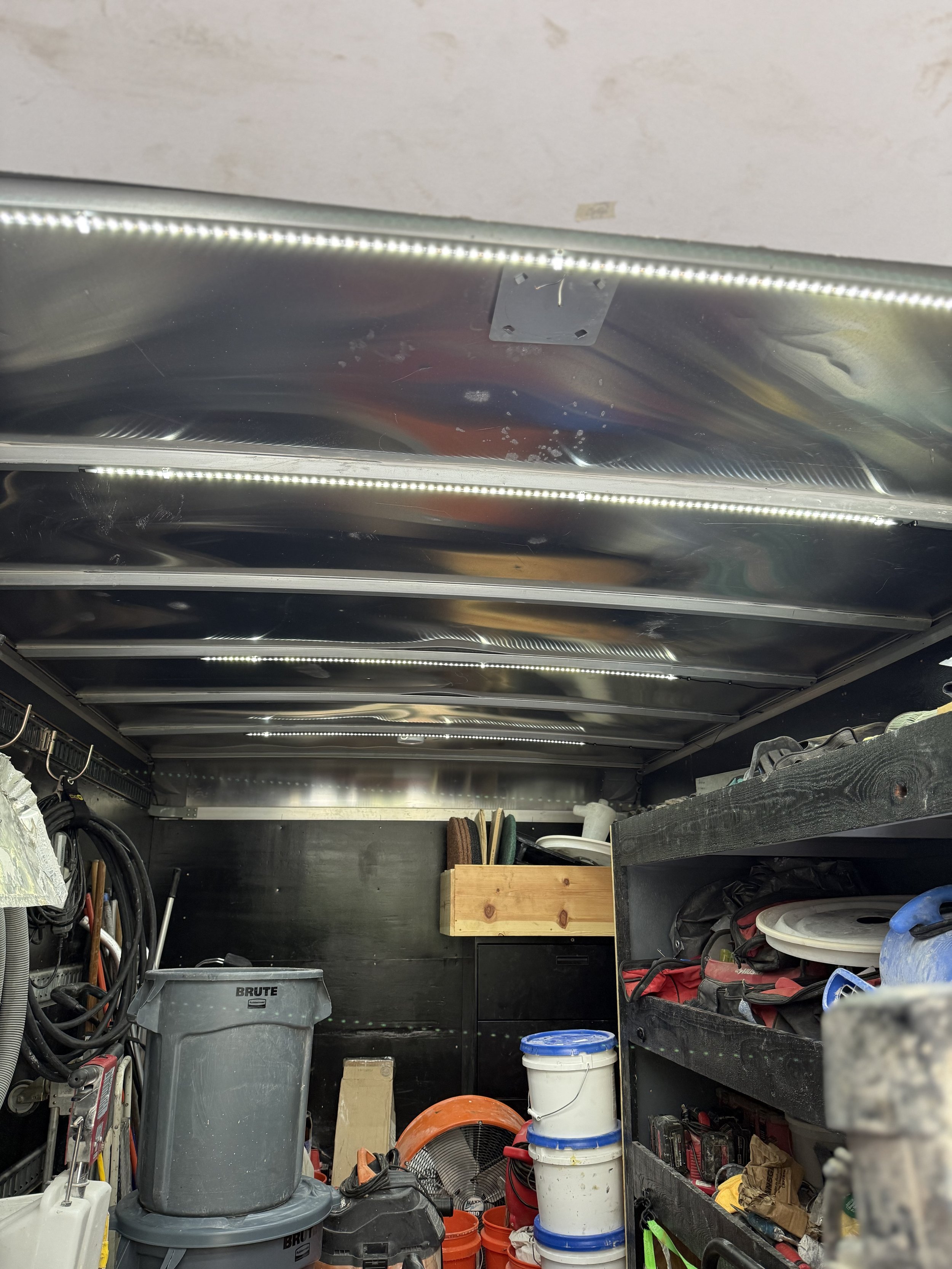 Inside a cluttered garage or workshop with shelves holding tools, supplies, and buckets. The ceiling has shiny metal panels with built-in LED strip lights.