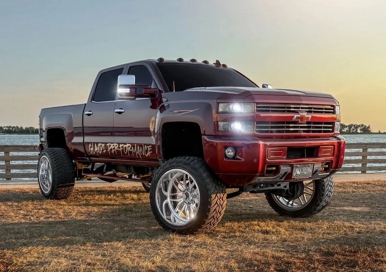 Red and black customized Chevrolet Silverado pickup truck with large tires, lifted suspension, and 'Club Performance' decal on the side, parked on a grassy area near a body of water at sunset.