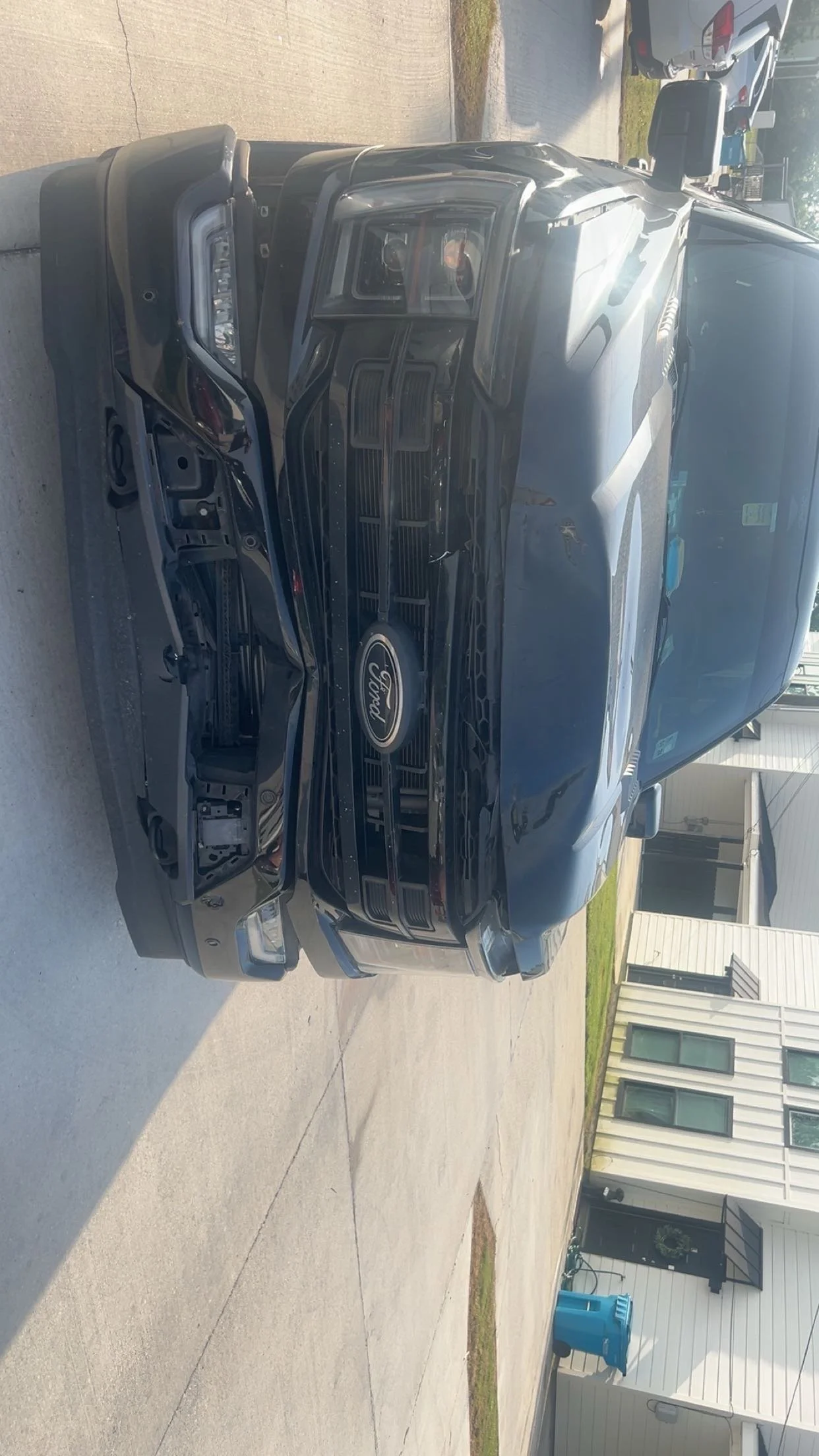 Front view of a black Ford F-150 truck with damaged front bumper, missing grille, and exposed internal components, parked on a driveway in front of a house.