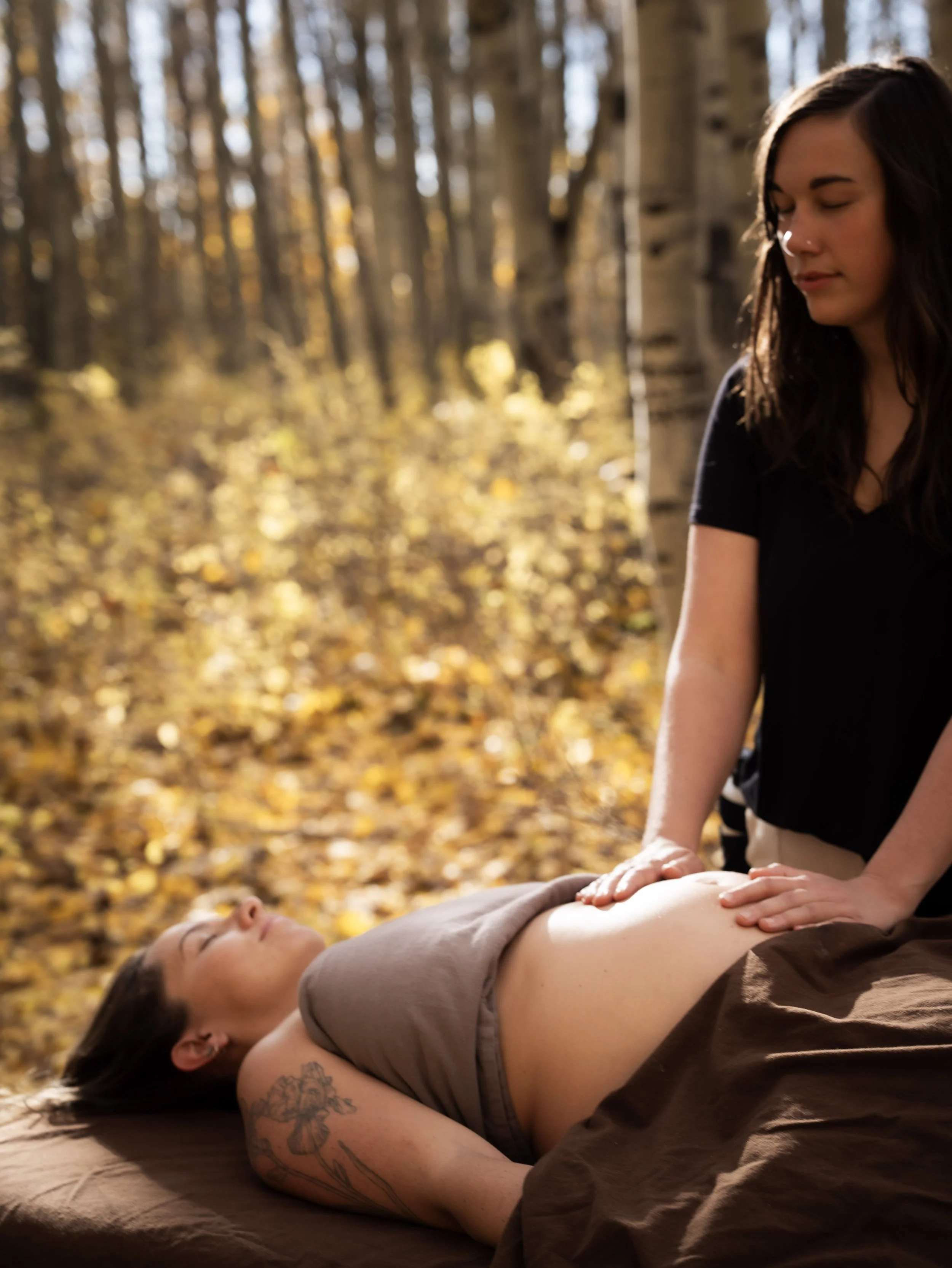 A woman receives a massage outdoors in a forest setting during autumn. She is lying on a massage table with her eyes closed, while another woman stands beside her, applying pressure to her stomach area.
