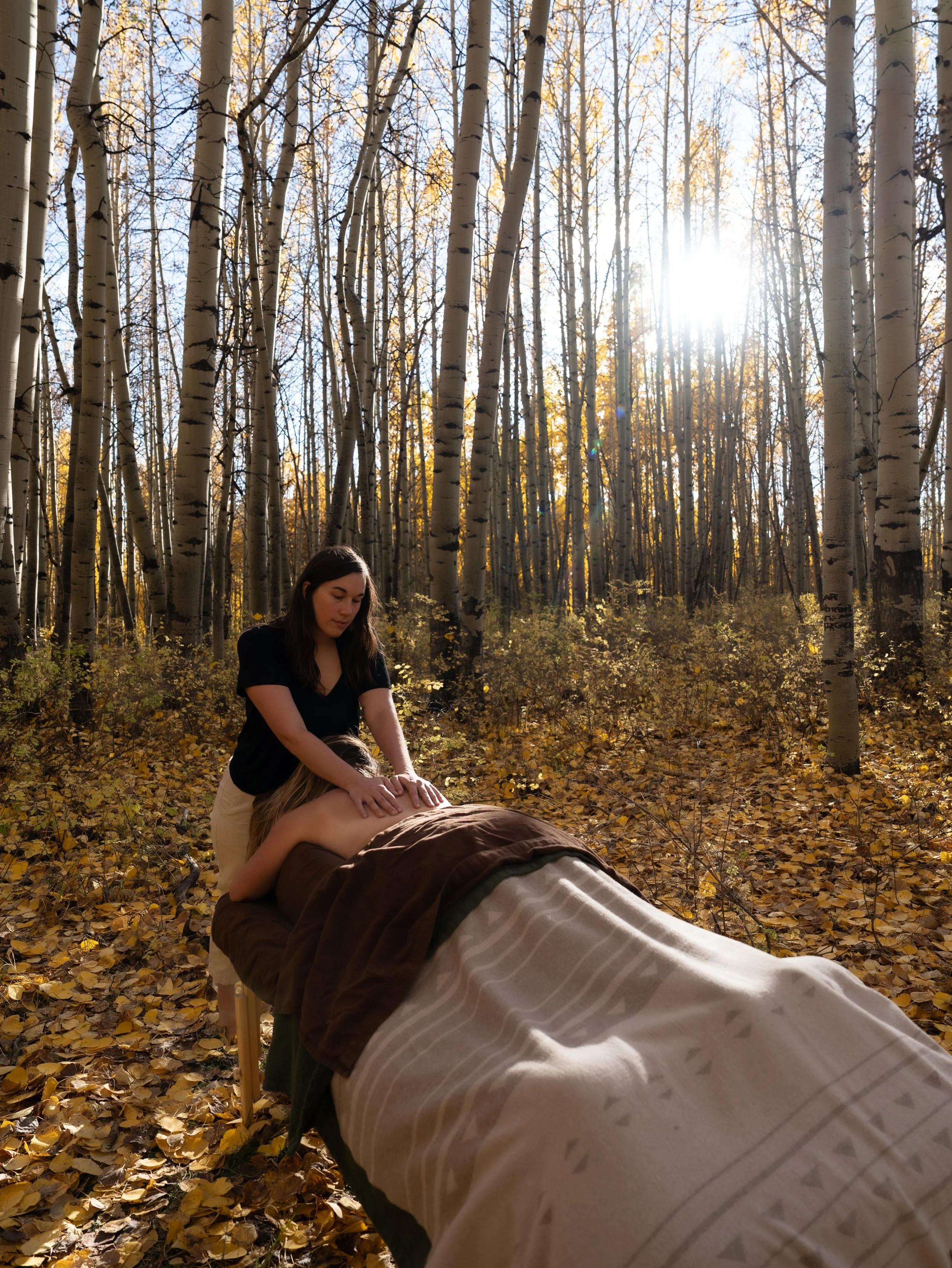 A woman is receiving a massage outdoors in a forest during autumn, with leaves on the ground and sunlight filtering through trees.