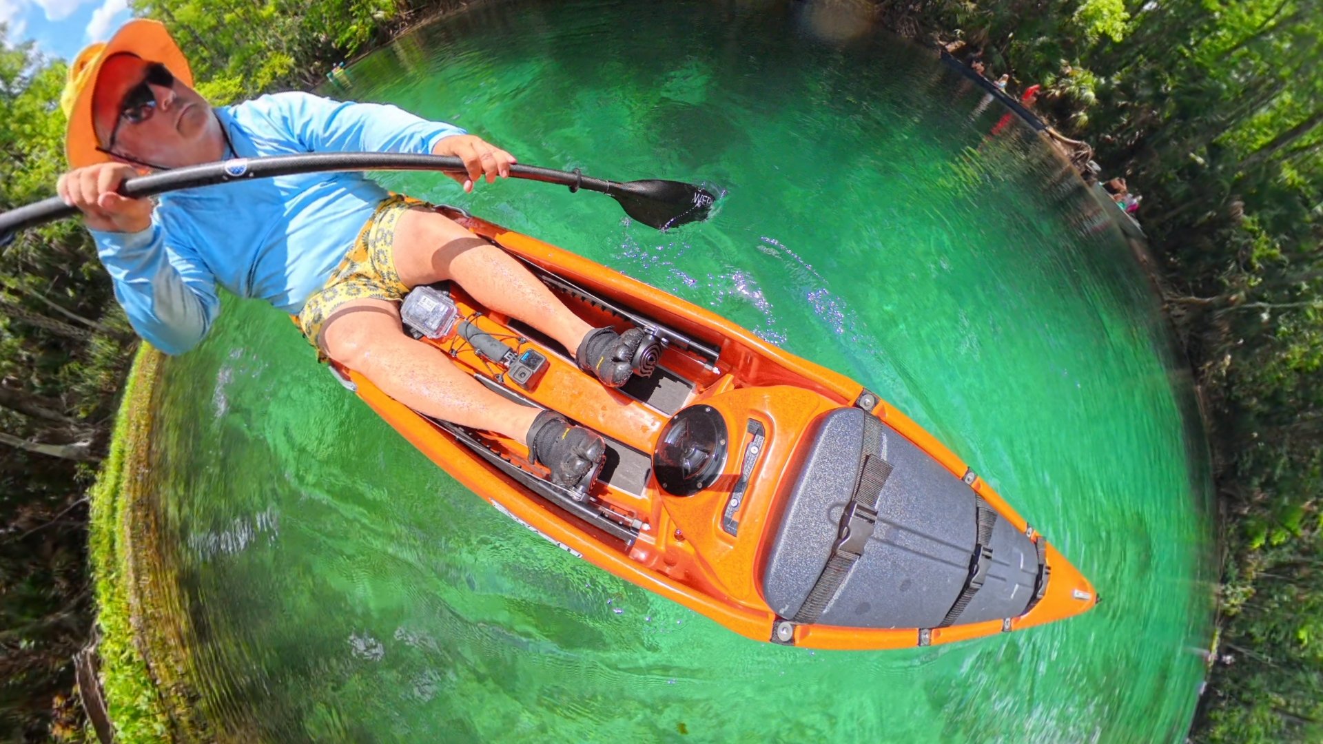 Person wearing a yellow hat, sunglasses, blue shirt, and floral shorts paddling an orange kayak on a clear, green water river surrounded by lush greenery.
