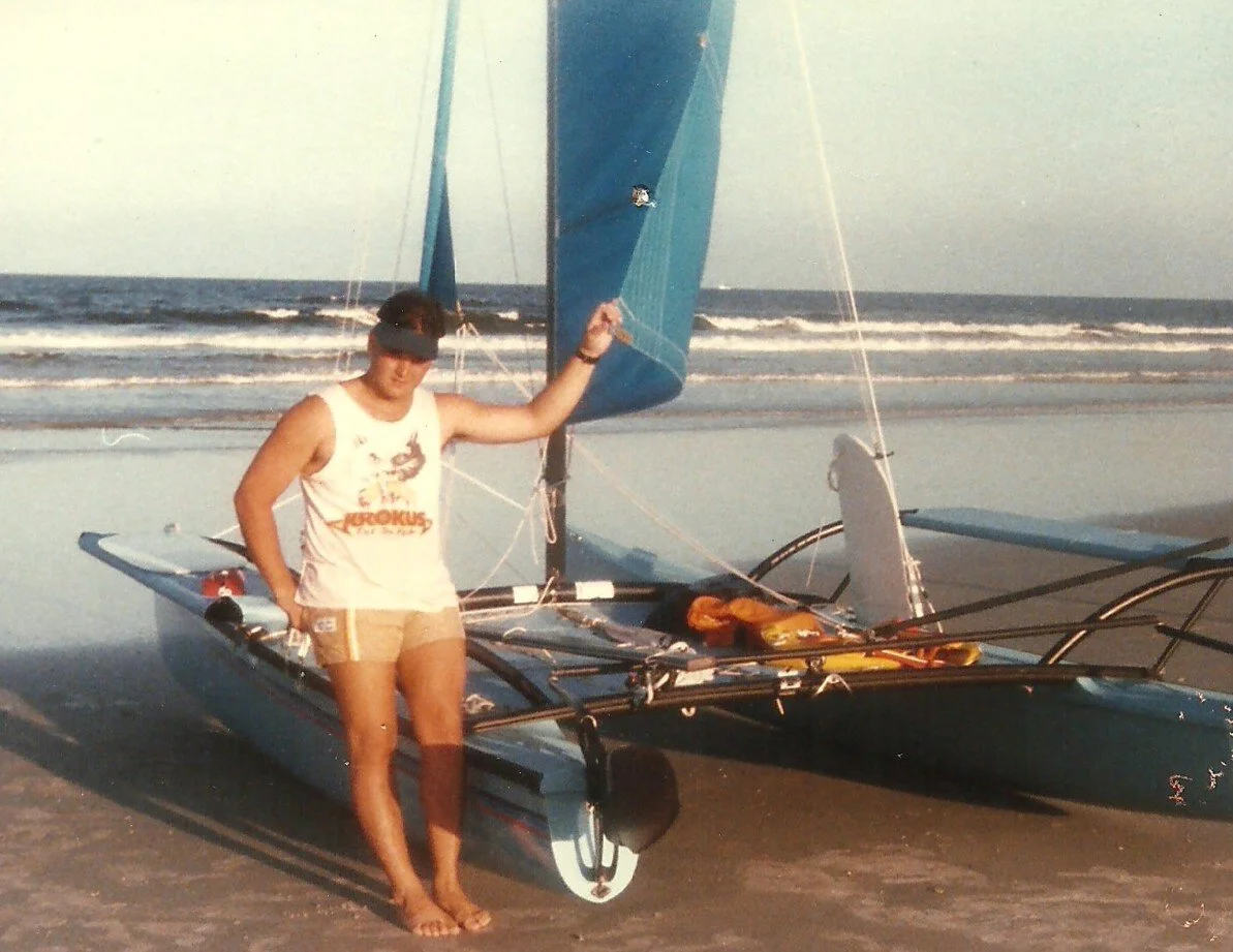 A young woman in a tank top, shorts, and a visor stands barefoot on a sandy beach next to a small sailing boat. The boat is on the sand with its sail partially raised, and the ocean with gentle waves is in the background.