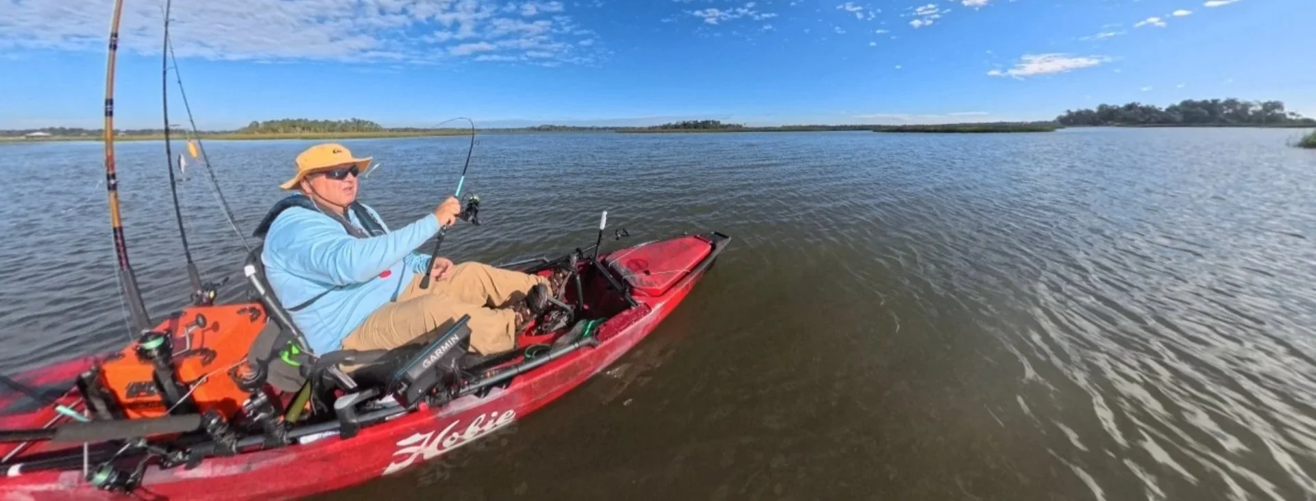 A man in a yellow wide-brim hat and sunglasses is sitting in a red kayak on a calm body of water, holding a fishing rod with a fish on the line. The sky is blue with some clouds, and there are trees on the distant shoreline.