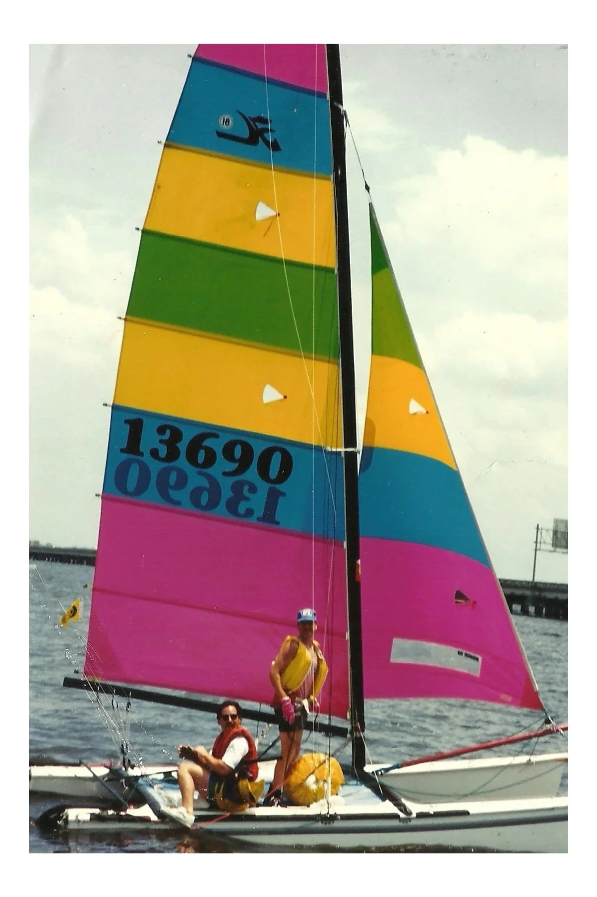 A colorful sailing boat with pink, yellow, green, blue, and black sails on the water, with two people on board, one sitting and one standing, under cloudy skies.