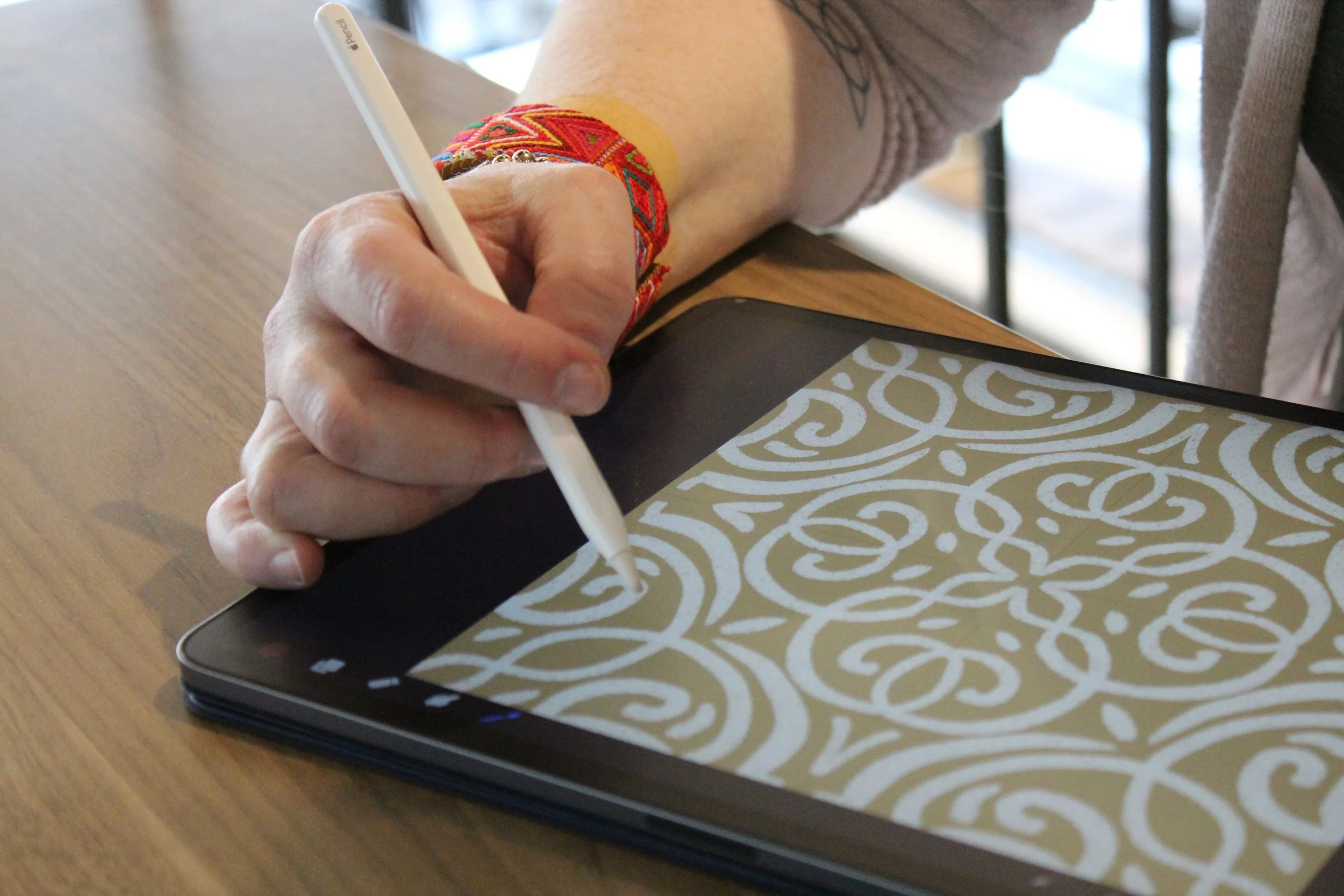 Person sitting at a wooden table drawing a decorative gold and white pattern on a tablet with a stylus, wearing a colorful bangle bracelet.